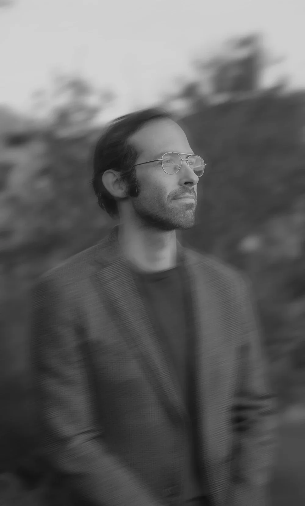 Black and white portrait of a man with glasses, in profile, outdoors with blurred trees in the background.