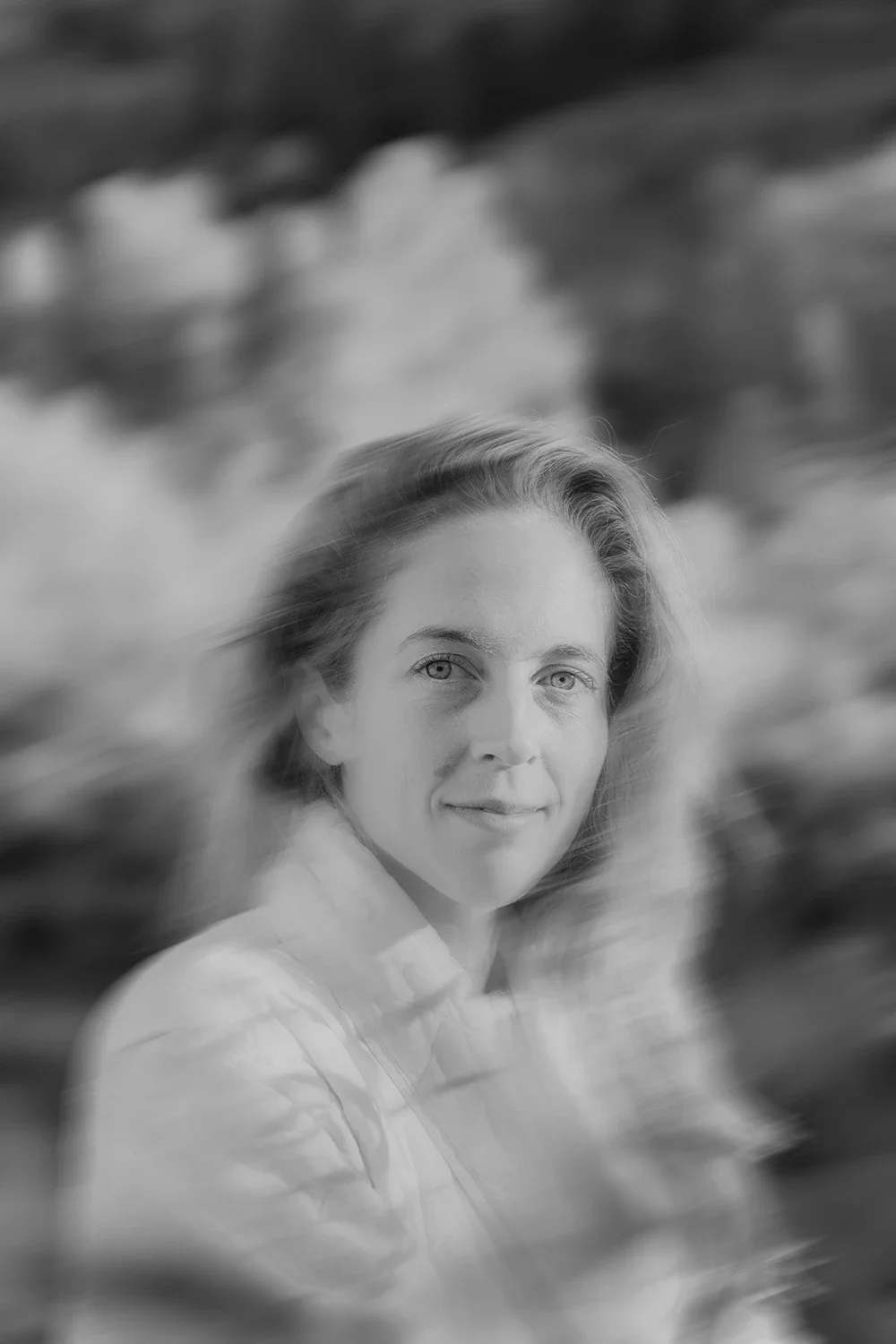 Black and white portrait of a woman with light skin and wavy hair, outdoors, with a blurred natural background.