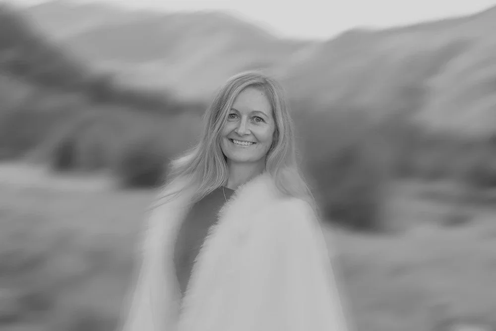 A woman with long blonde hair smiling outdoors with a blurred landscape of rolling hills and cloudy sky in the background