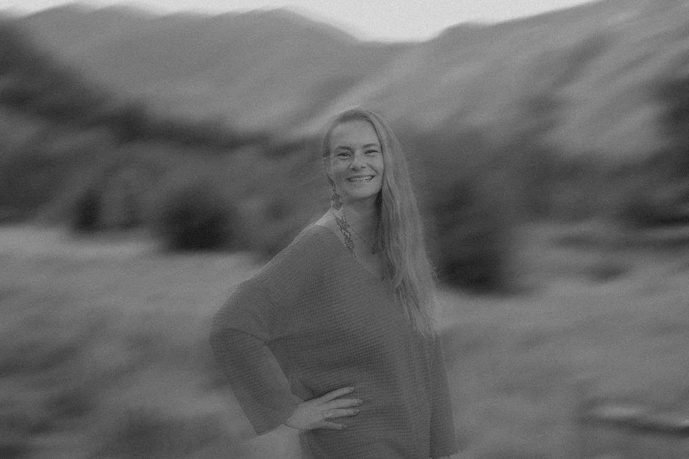 A smiling woman with long hair standing outdoors in a natural landscape with mountains in the background, wearing a loose sweater.