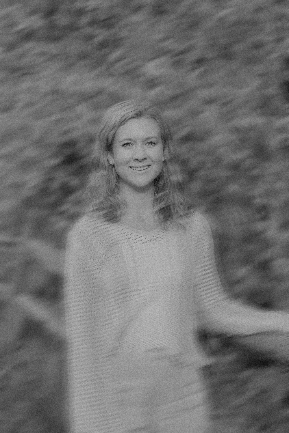Black and white photo of a woman smiling, standing outdoors near water with ripples.