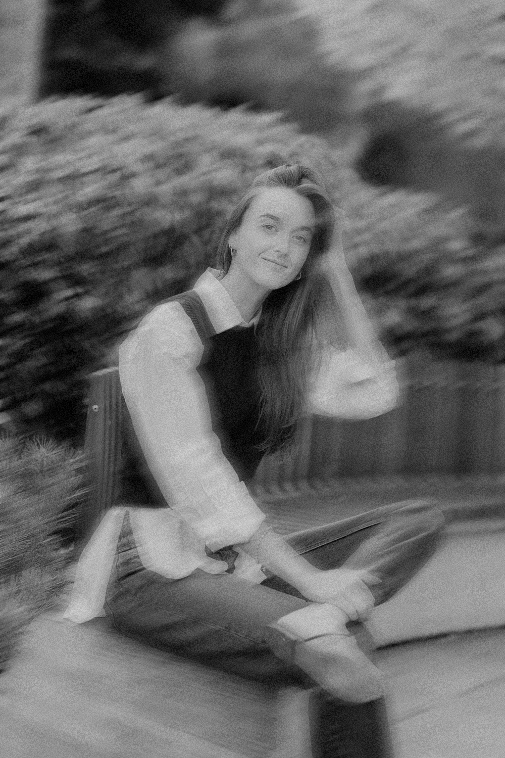 A woman with long hair sitting outdoors, smiling with her hand in her hair, wearing a white shirt and dark vest, with a blurred background.