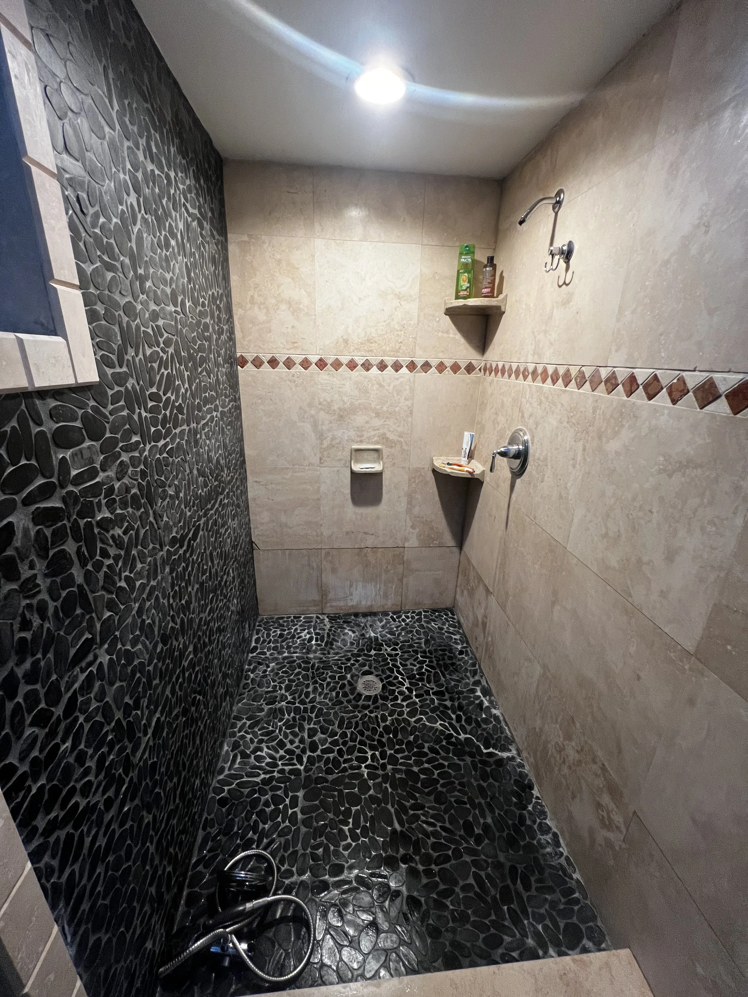 Empty shower with black pebble stone floor and dark pebble stone accent wall on the left, beige tiled walls, shelves with toiletries, and a single overhead light.