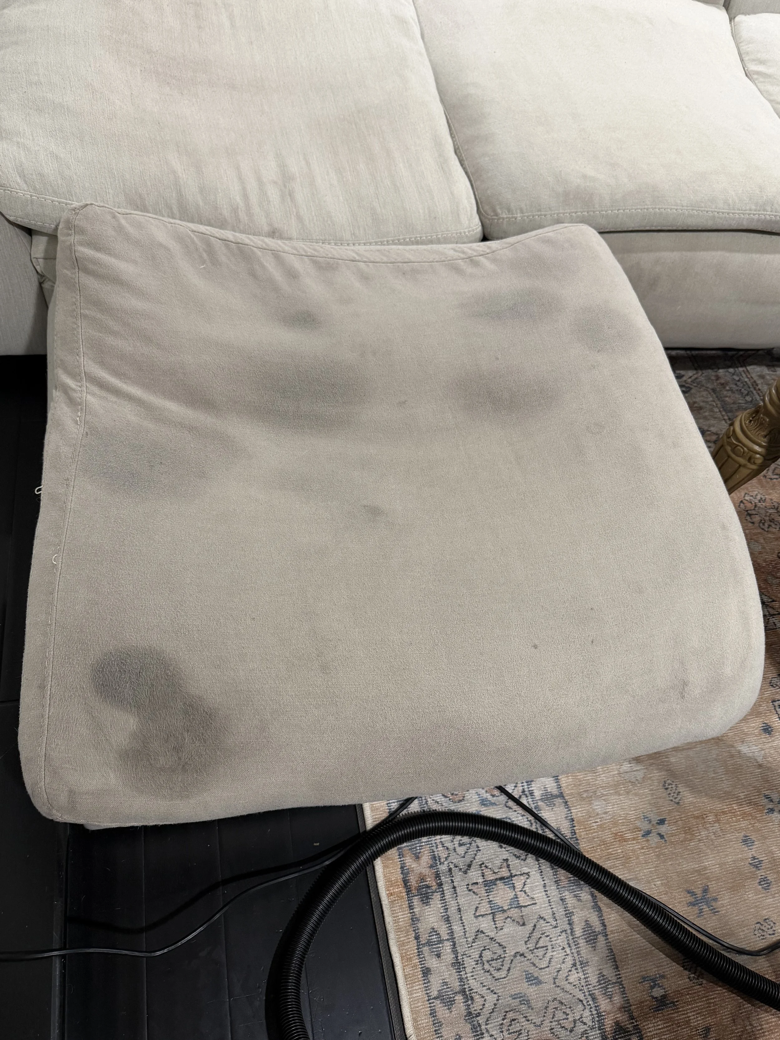A beige ottoman with multiple dark stains on the seat, situated on a patterned area rug near a cream-colored sofa.