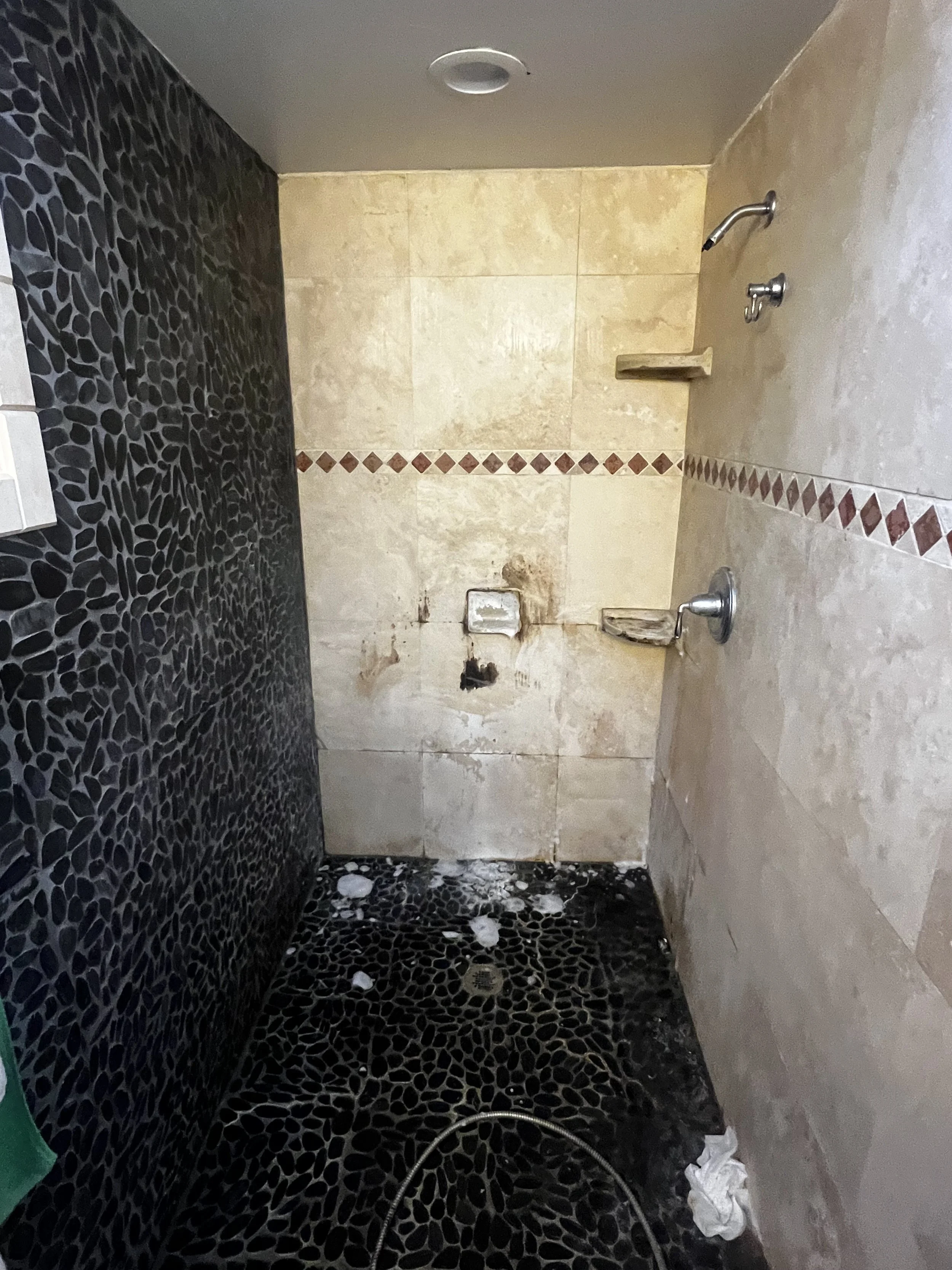 A shower area under renovation with black pebble tiles on the floor and a textured beige wall with a decorative border. No shower fixtures installed, and debris on the floor.