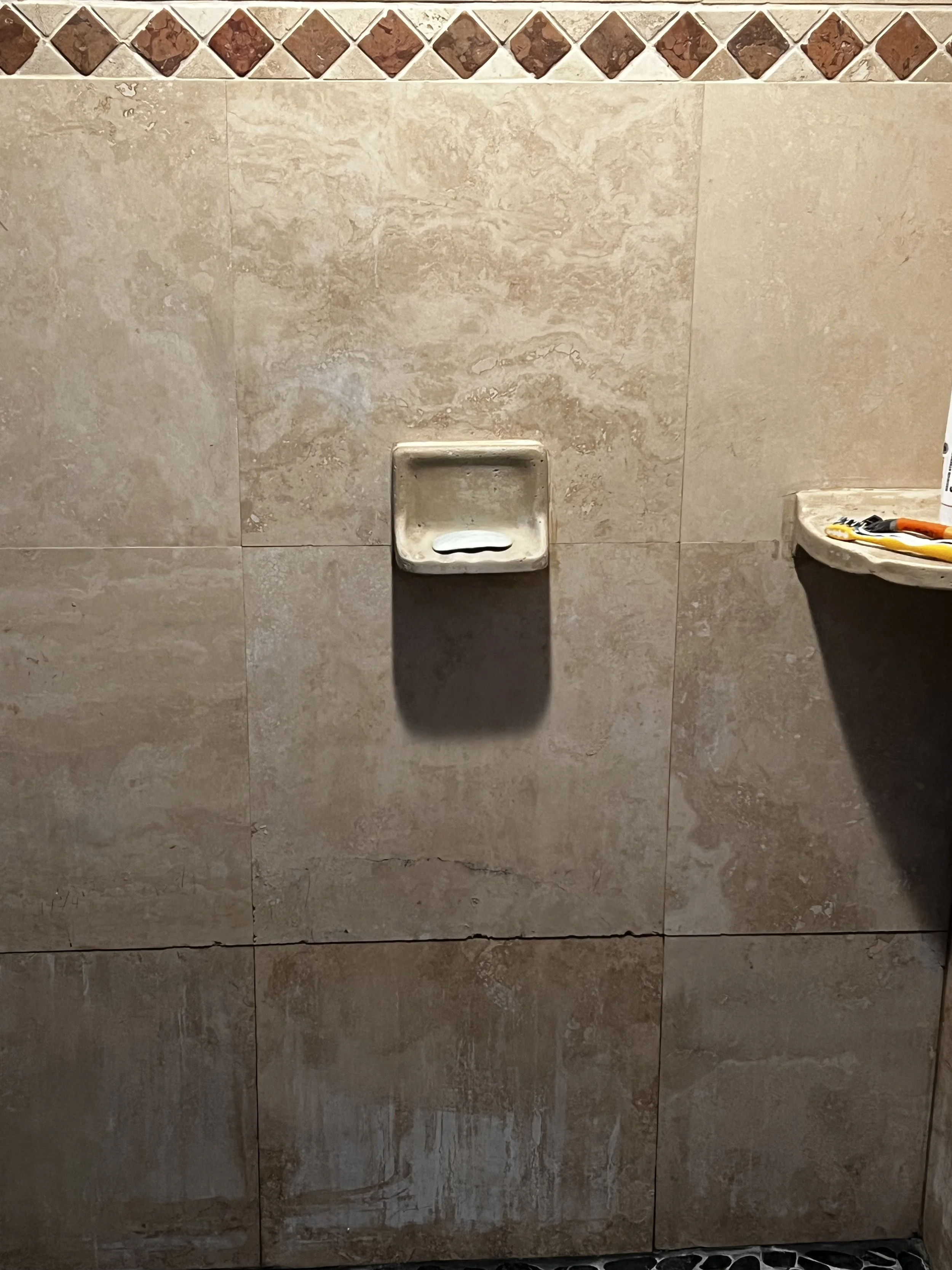 A wall in a shower with beige tiles, a small built-in soap dish, and a ledge with tools on it.