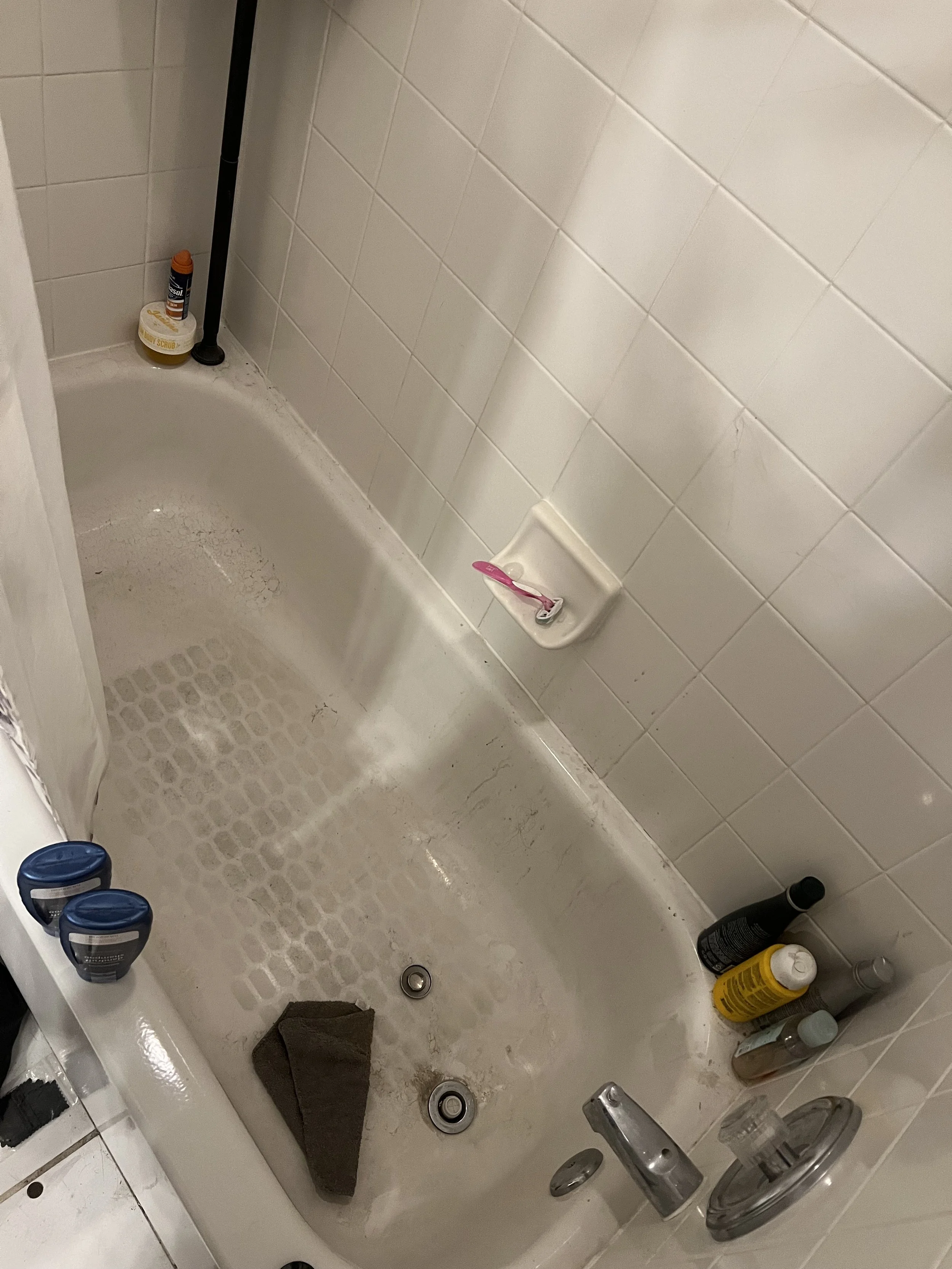 Dirty bathtub with various bottles and a brown cloth, surrounded by white tiled walls with a toothbrush in a soap dish.