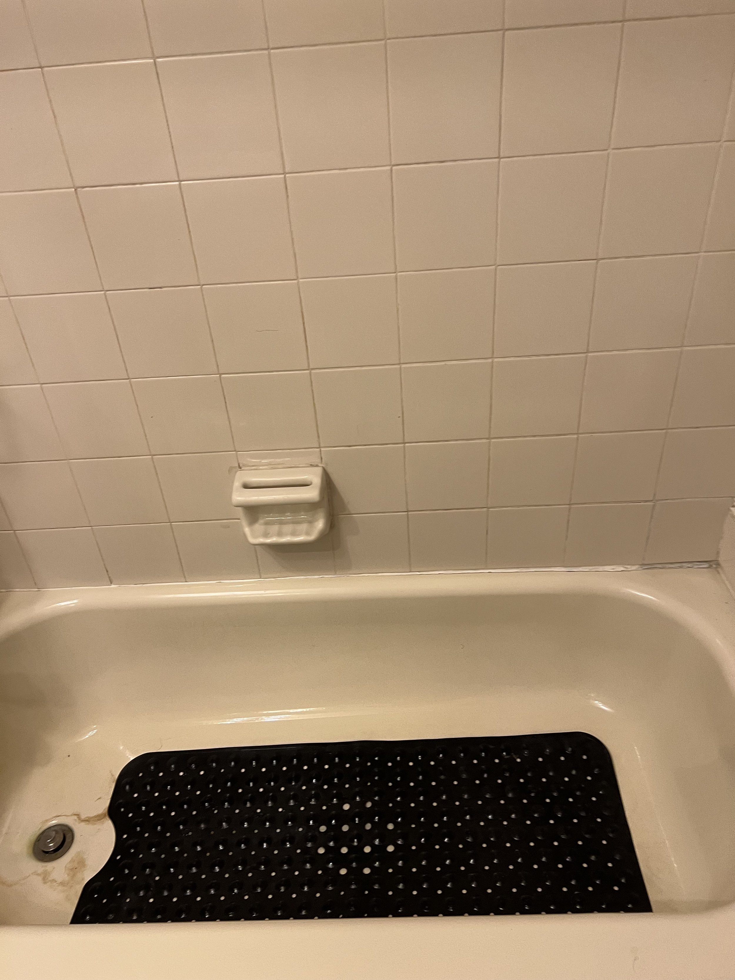 Bathroom bathtub with a black rubber mat and white tiled wall, soap dish attached to the wall.