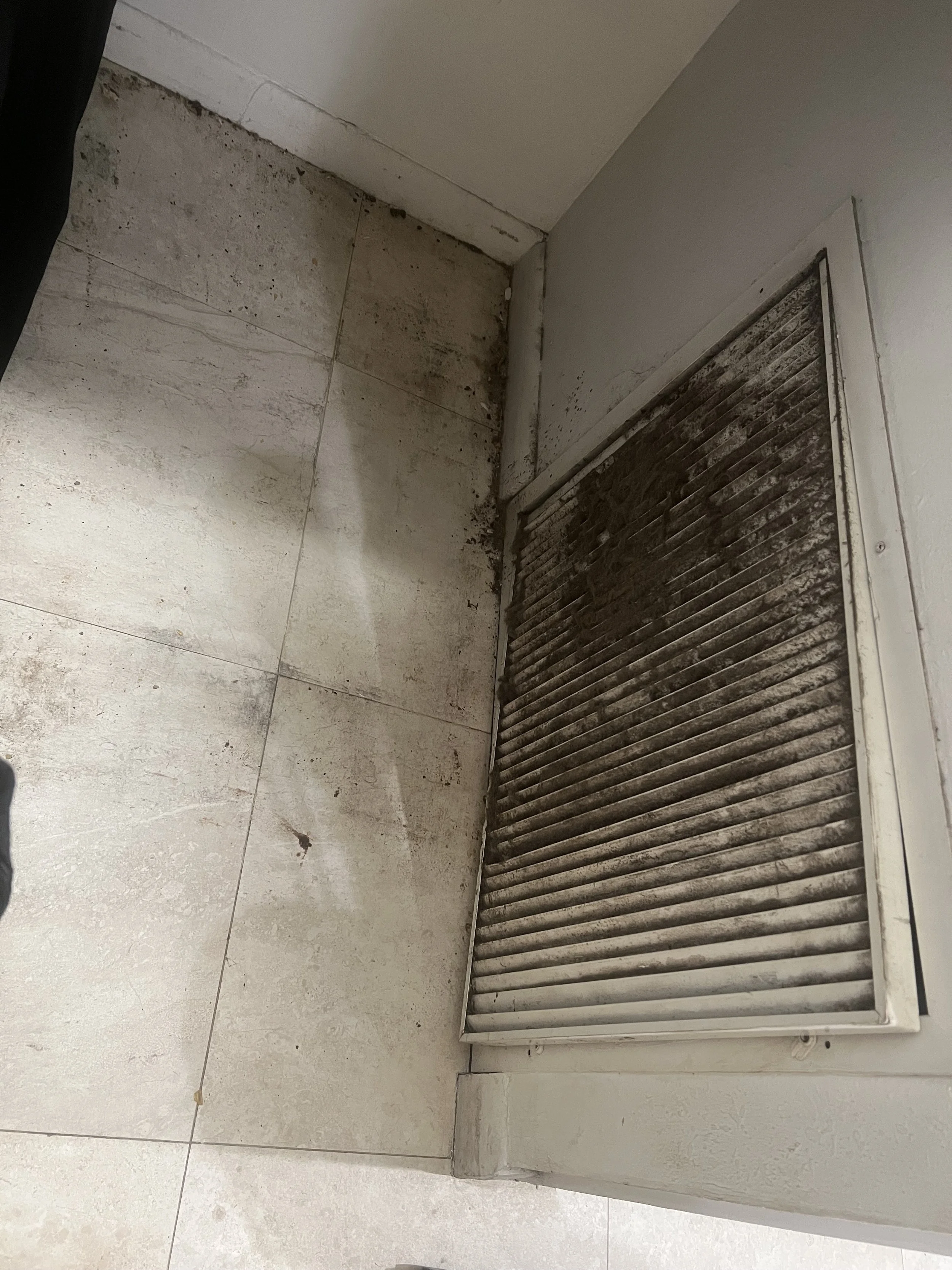 A dirty air vent cover on a tiled floor near a wall, with visible dust and dirt buildup.