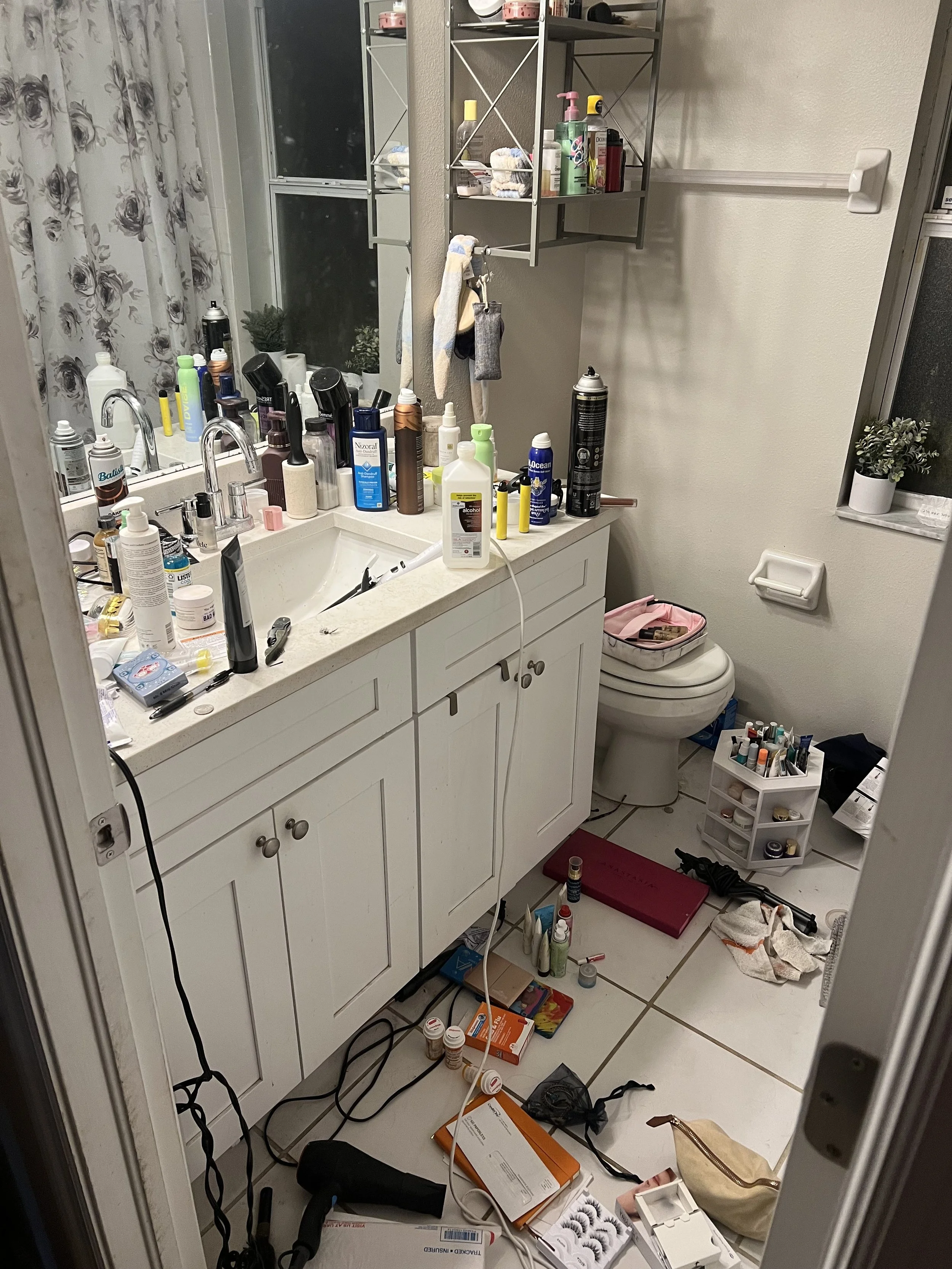 Messy bathroom with cluttered sink, toiletries, hair tools, and scattered items on the floor including a hairdryer, curling iron, and makeup supplies.