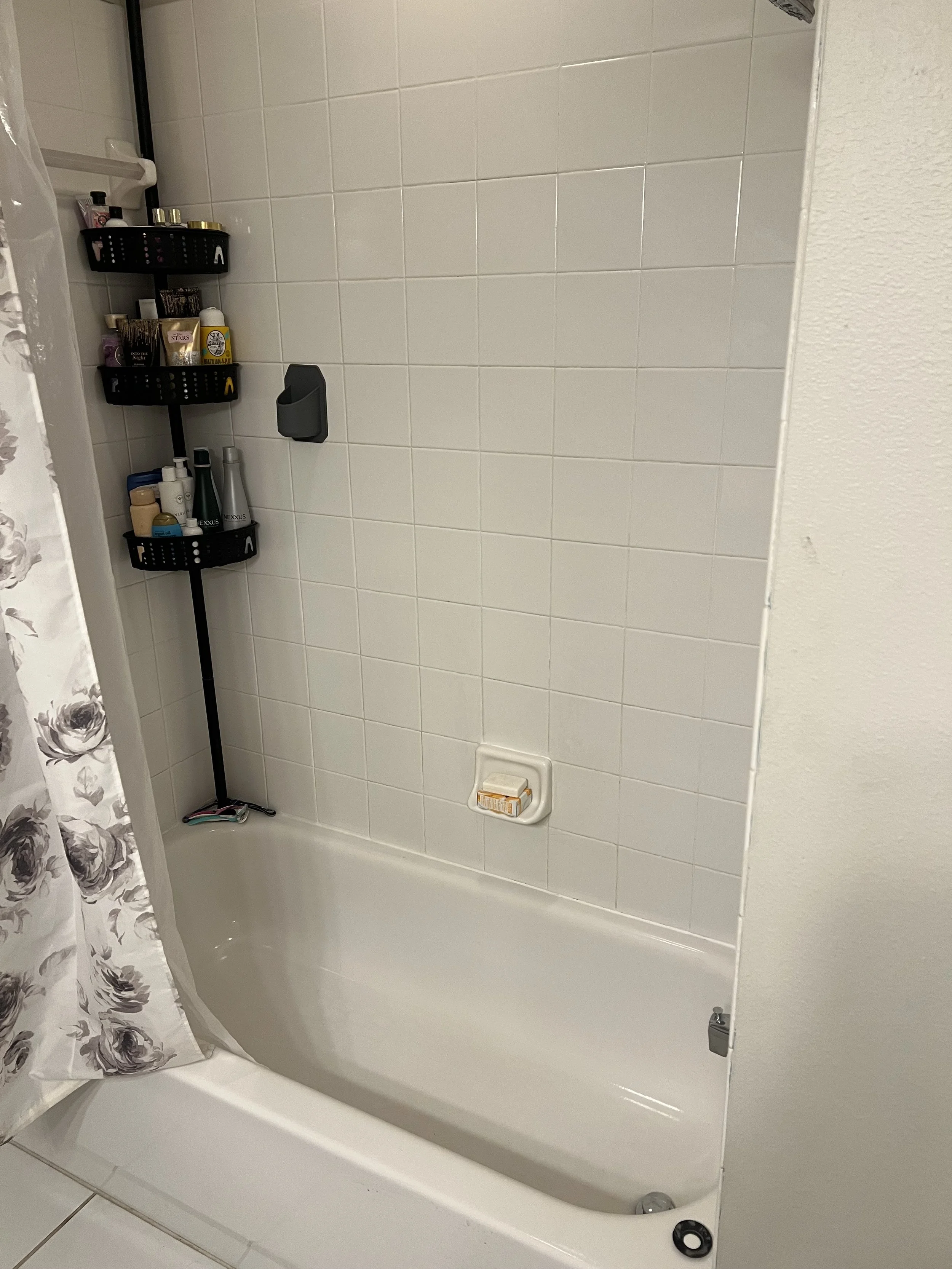 Empty bathtub with a shower curtain on the left side, a wall-mounted soap holder with soap, and a black caddy on a vertical rod holding various bottles and toiletries in a tiled bathroom.