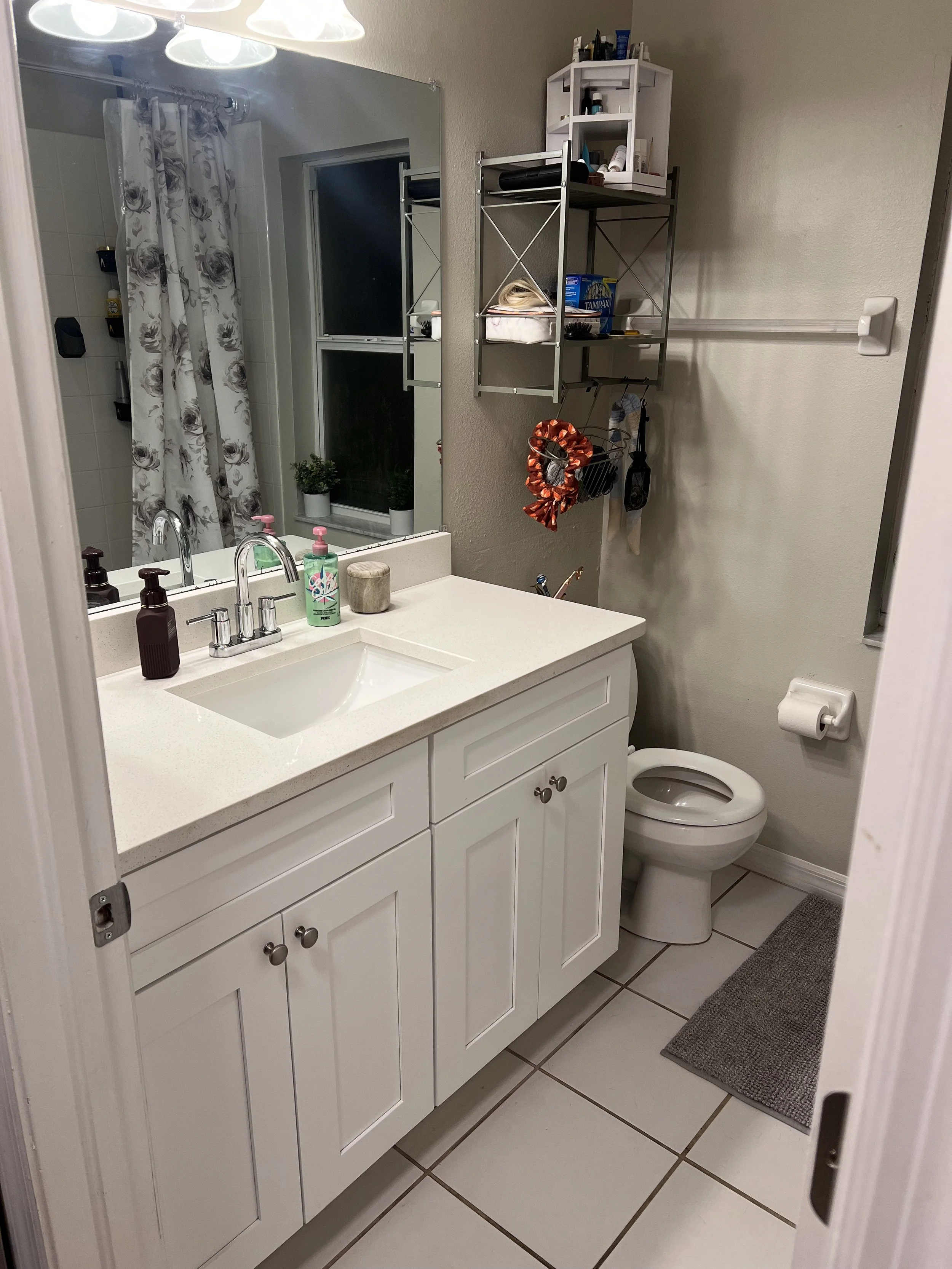 A small bathroom with a white vanity, a mirror, and a sink. On the counter are soap dispensers and a candle. There is a toilet with a toilet paper roll on the wall next to it. Above the toilet, a wall-mounted shelf holds various toiletries. A gray ru
