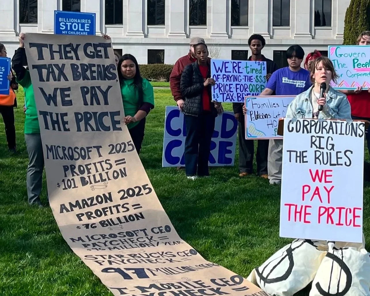 Hannah Lindell-Smith of the Washington Bus speaks at the Power Building Table's media event in support of a Millionaires' Tax. She stands at a podium next to a long list of corporation profits.
