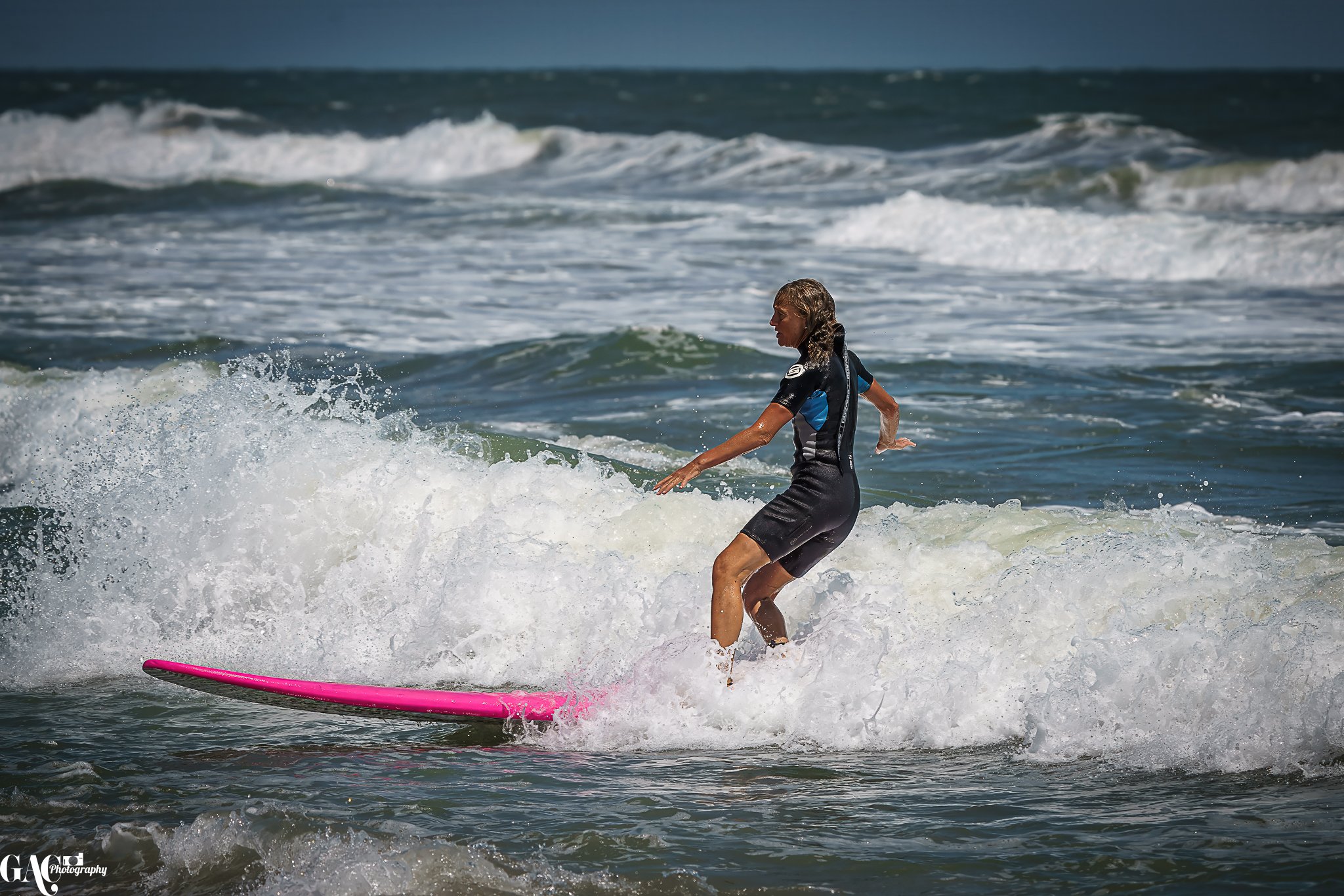 A woman surfing on a pink surfboard in the ocean with white waves in the background.