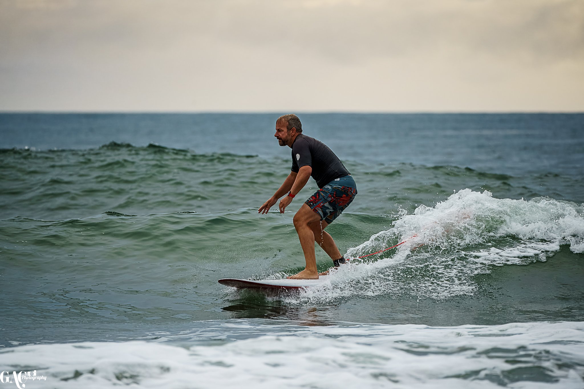 A man surfing on a small wave in the ocean, wearing a black shirt and multicolored swim shorts, with a cloudy sky above.