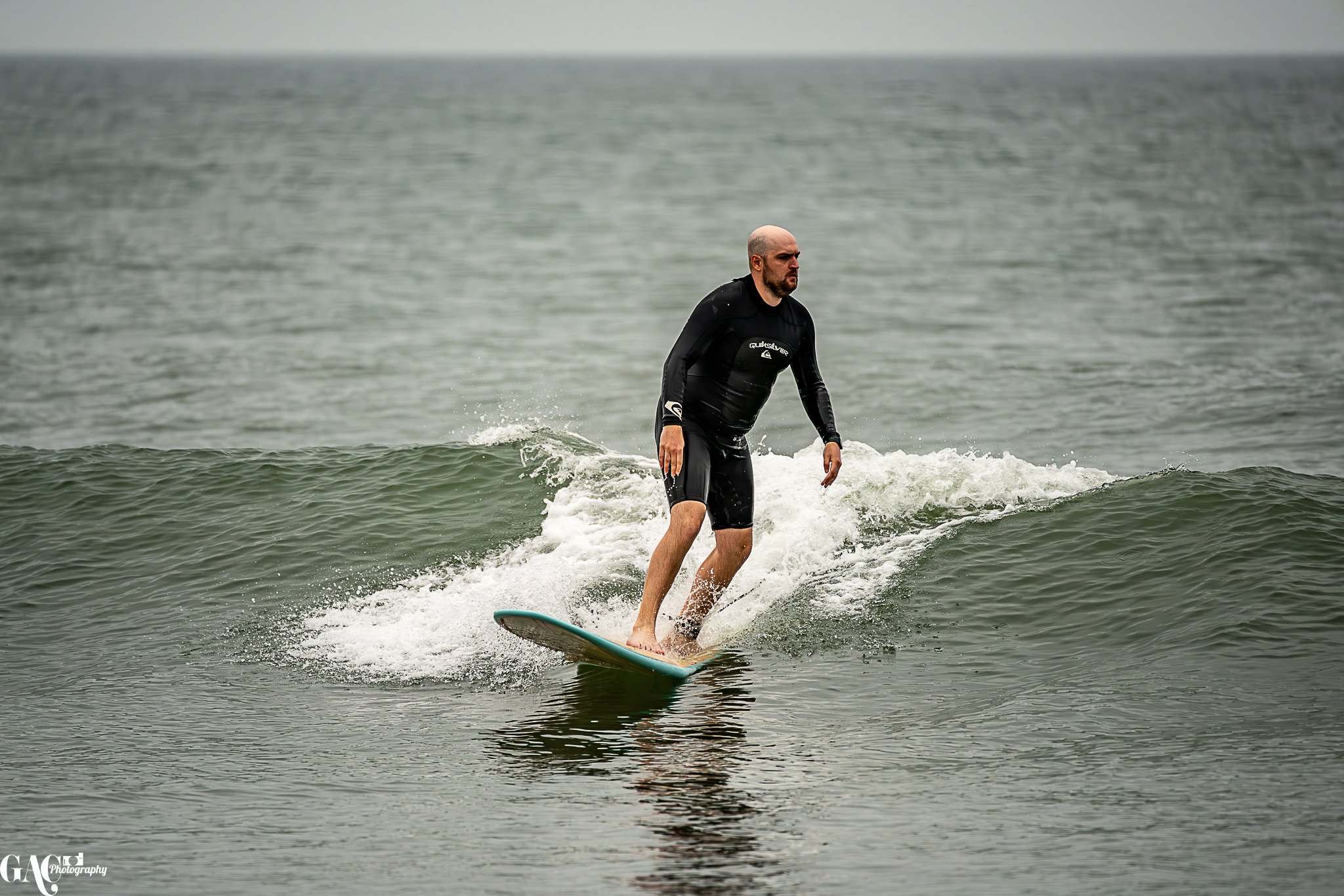 A man surfing on a small wave in the ocean, wearing a black wetsuit with a bald head and beard.
