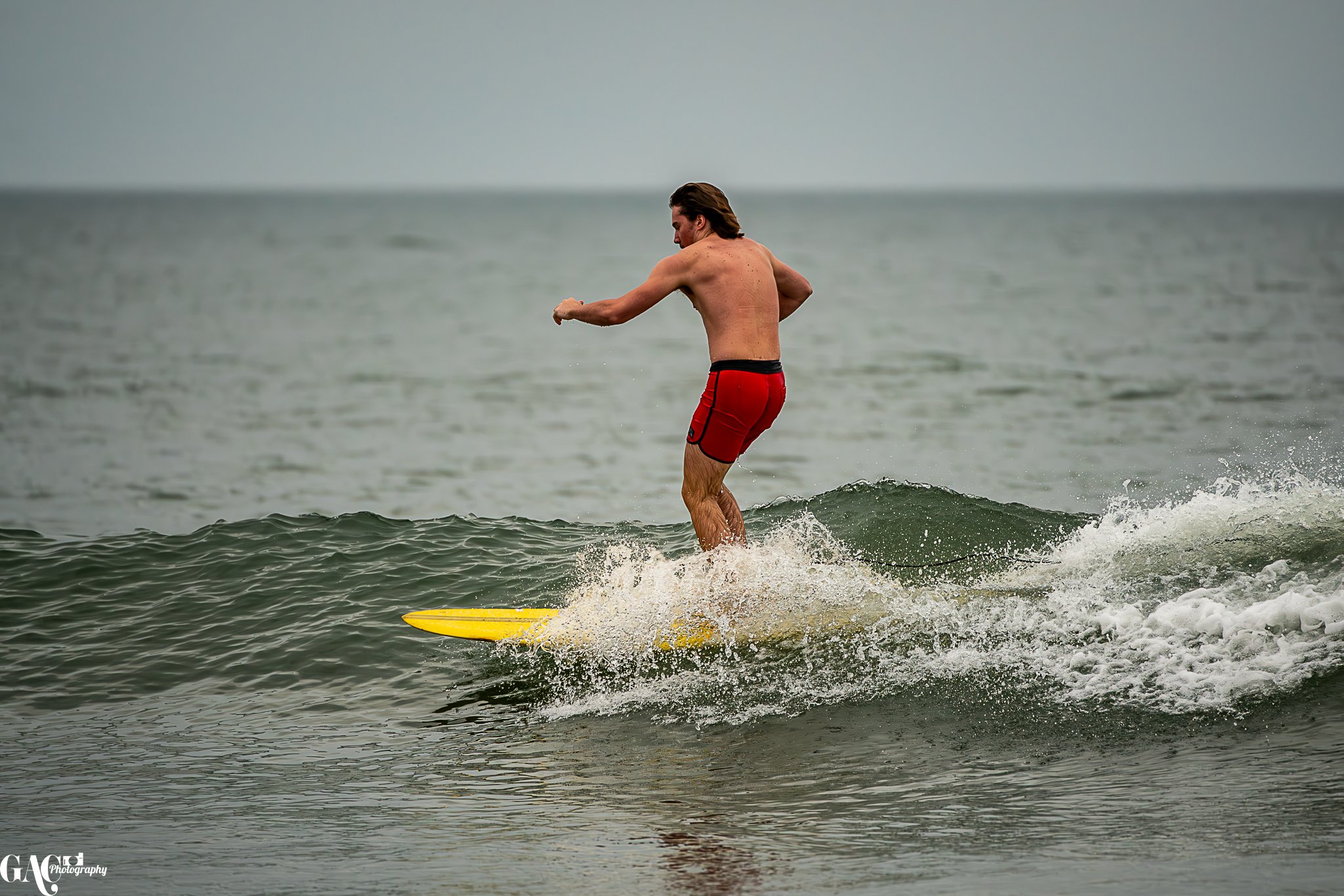A man surfing on a yellow surfboard in the ocean, wearing red shorts, with gray sky and water in the background.