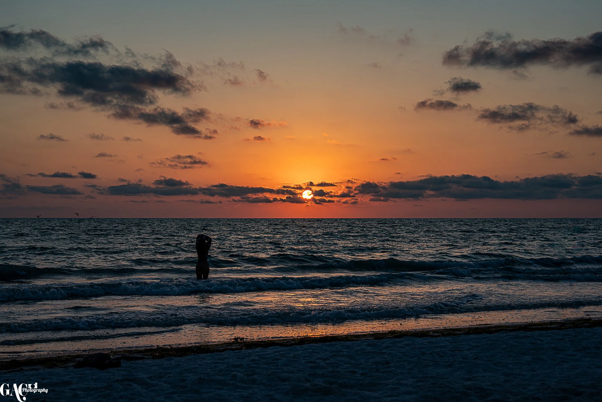 A person standing in the ocean at sunset with clouds in the sky.