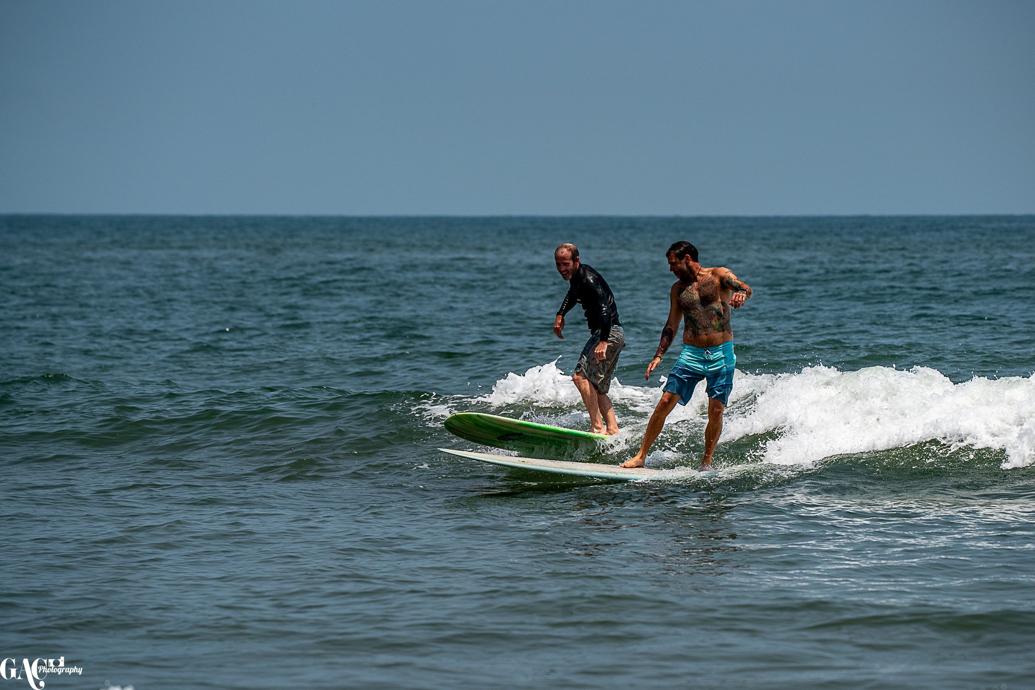 Two men surfing on a small wave in the ocean, one with tattoos and the other wearing a black wetsuit.