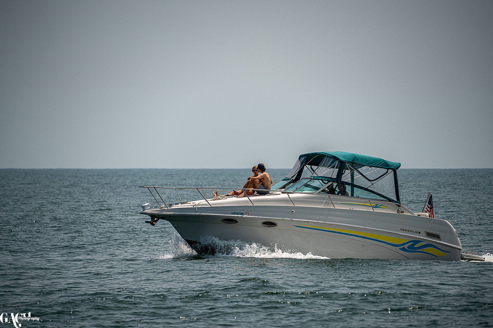 A white motorboat with a green canopy cruising on the water, with two people sitting on the deck, one wearing sunglasses.