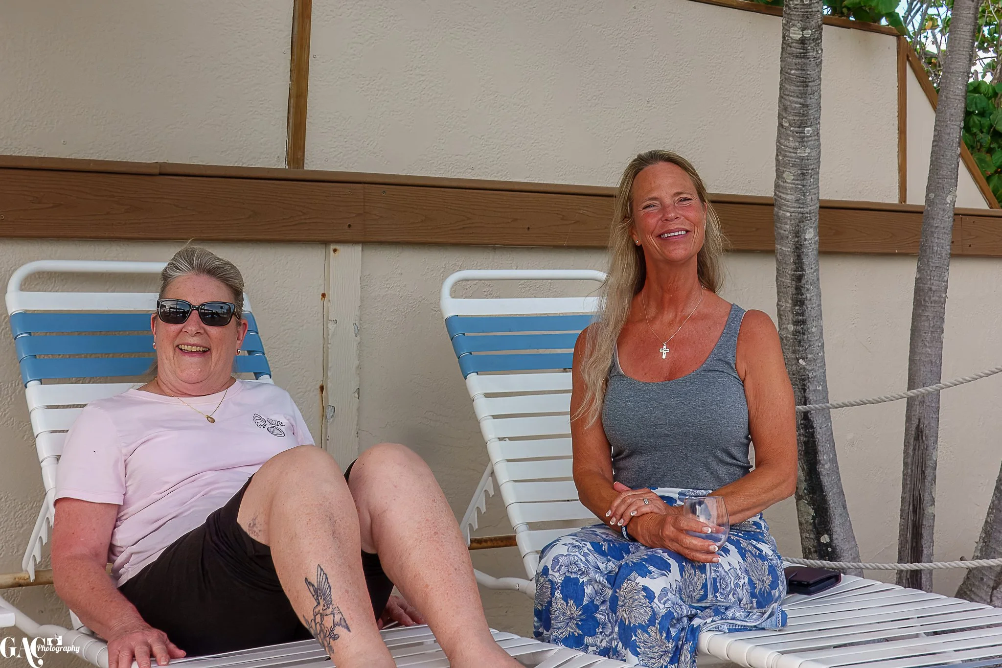 Two women sitting on lounge chairs, one wearing sunglasses and a white shirt, and the other in a gray tank top with a floral skirt, smiling outdoors with palm trees in the background.