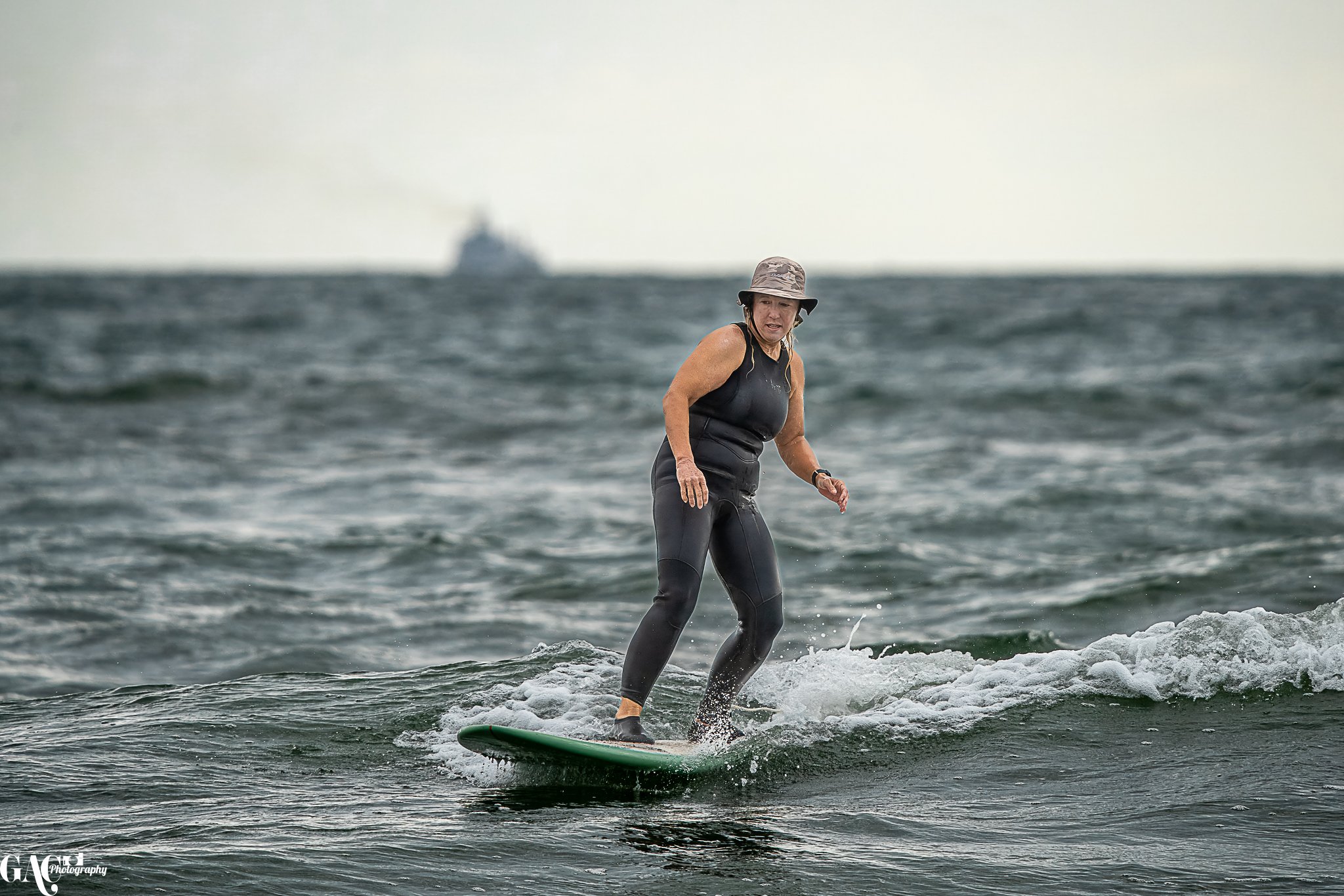 Woman wearing a black wetsuit and a hat riding a surfboard on a small wave in the ocean with a ship in the background.