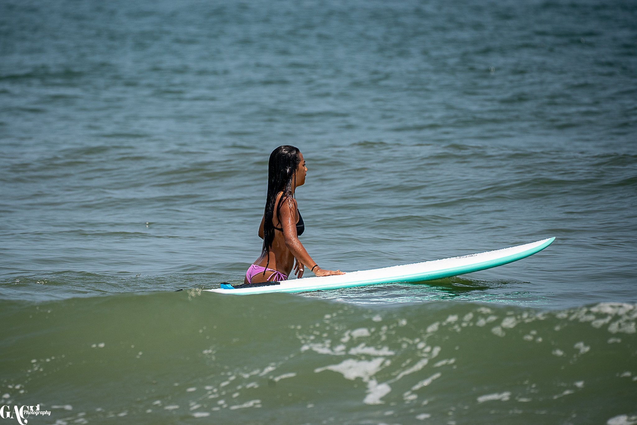 A woman with long dark hair in a black bikini top and purple bottoms, paddling a white surfboard in the ocean.