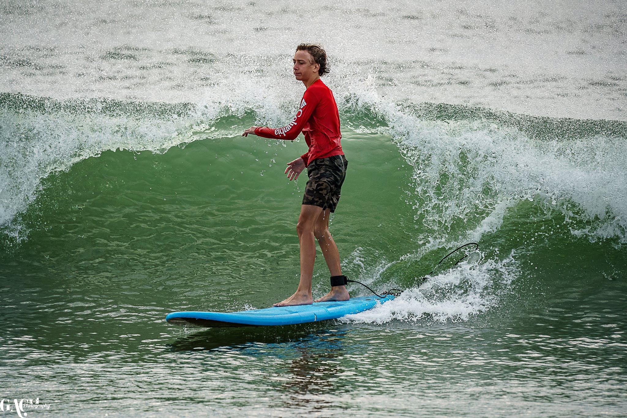 A young person surfing on a blue surfboard in the ocean, riding a green wave, wearing a red long-sleeve shirt and camouflage shorts.