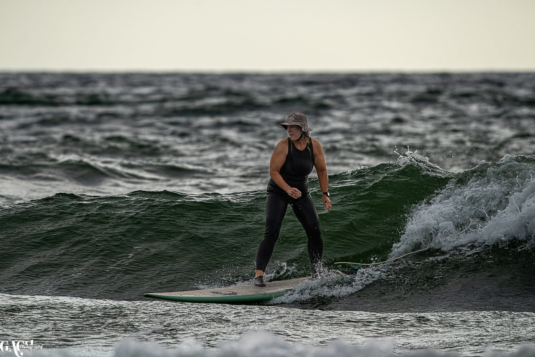 A woman wearing a black sleeveless top, black leggings, and a wide-brim hat is surfing on a green surfboard in the ocean.
