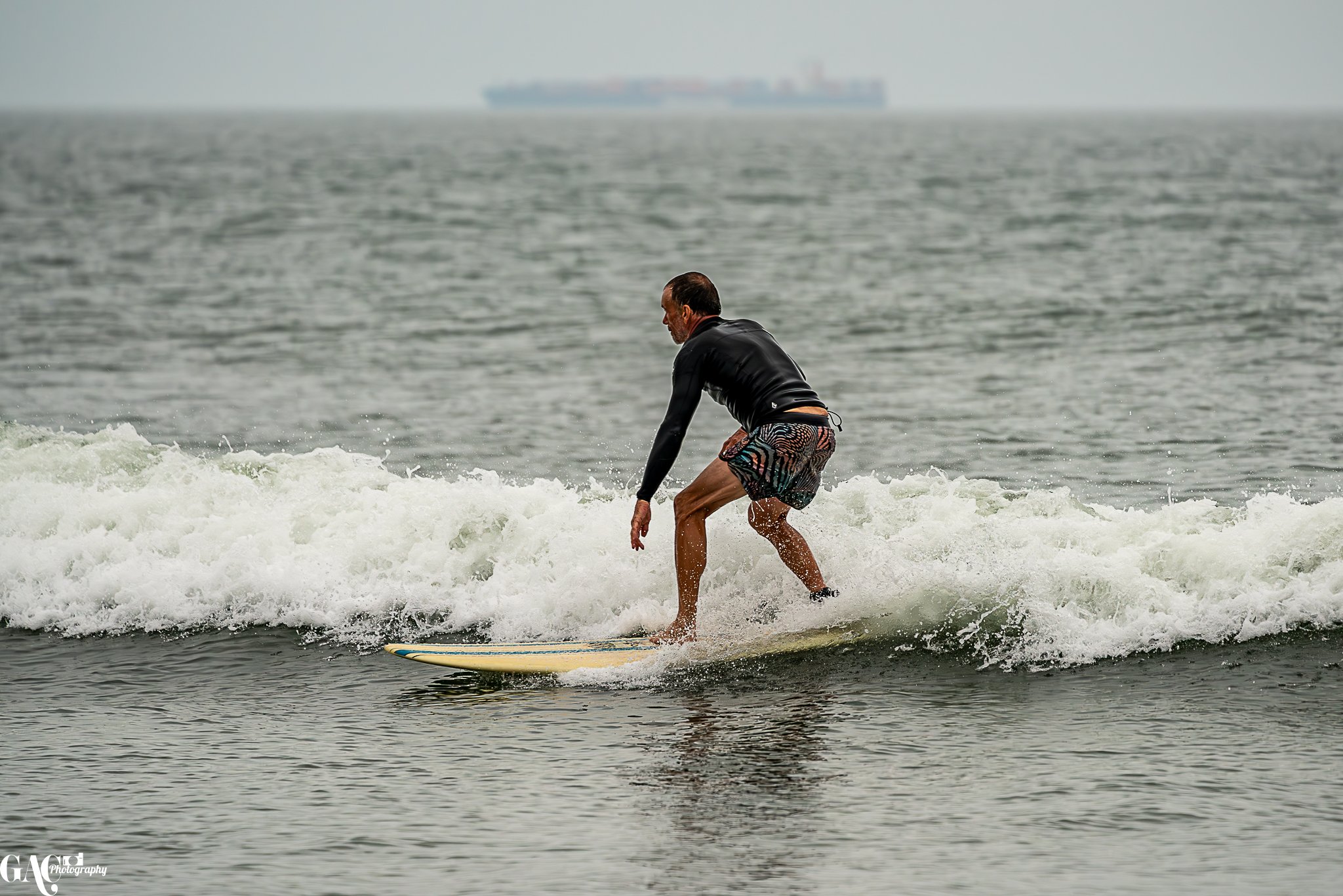 A man surfing on a small wave in the ocean, wearing a black rash guard and colorful swim shorts, with a large cargo ship in the background.