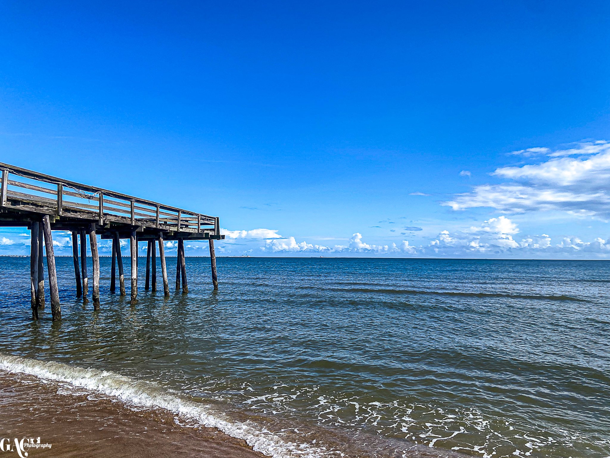 A wooden pier extends into the ocean with a sandy beach in the foreground and a blue sky with scattered clouds above.