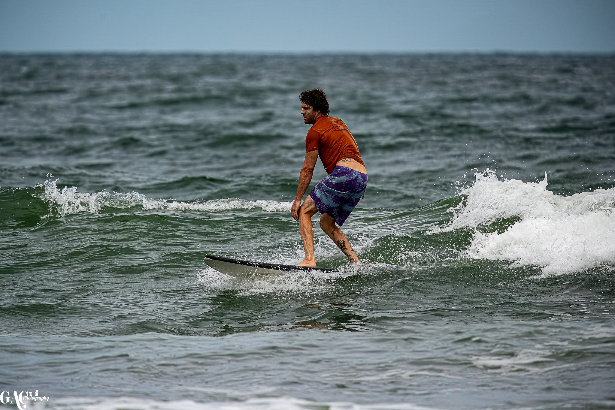 Man surfing on a wave in the ocean, wearing a brown t-shirt and purple patterned shorts.