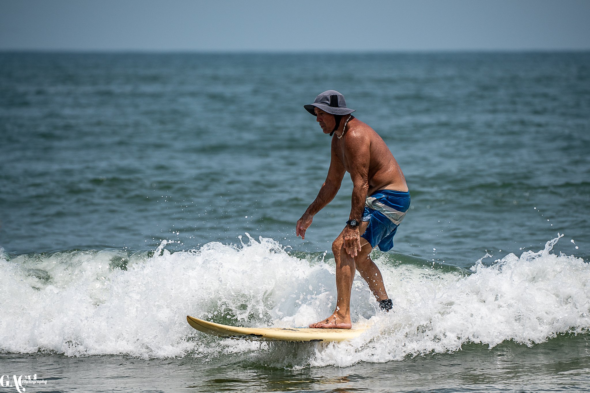 An elderly man wearing a hat and blue shorts is surfing on a wave at the beach.