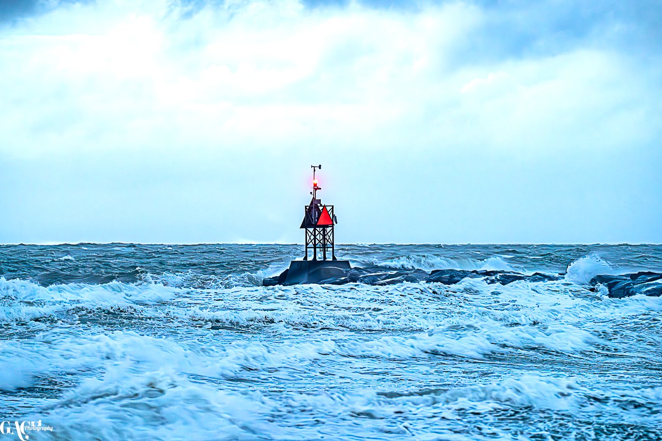 A lighthouse standing on rocks in a rough sea with crashing waves and a cloudy sky.