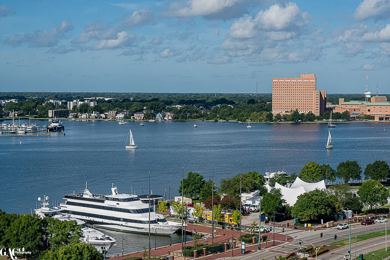 A scenic view of a harbor with sailboats and yachts, a waterfront area with boats docked, a large river or bay, buildings and trees in the background, and a partly cloudy sky.