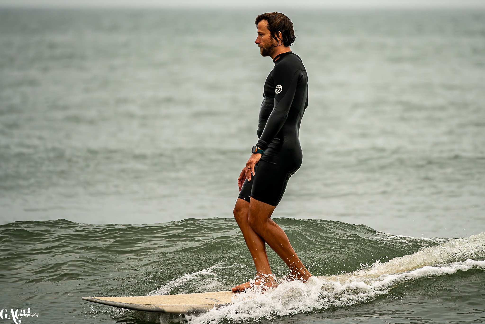 A man in a black wetsuit riding a surfboard on small waves in the ocean.