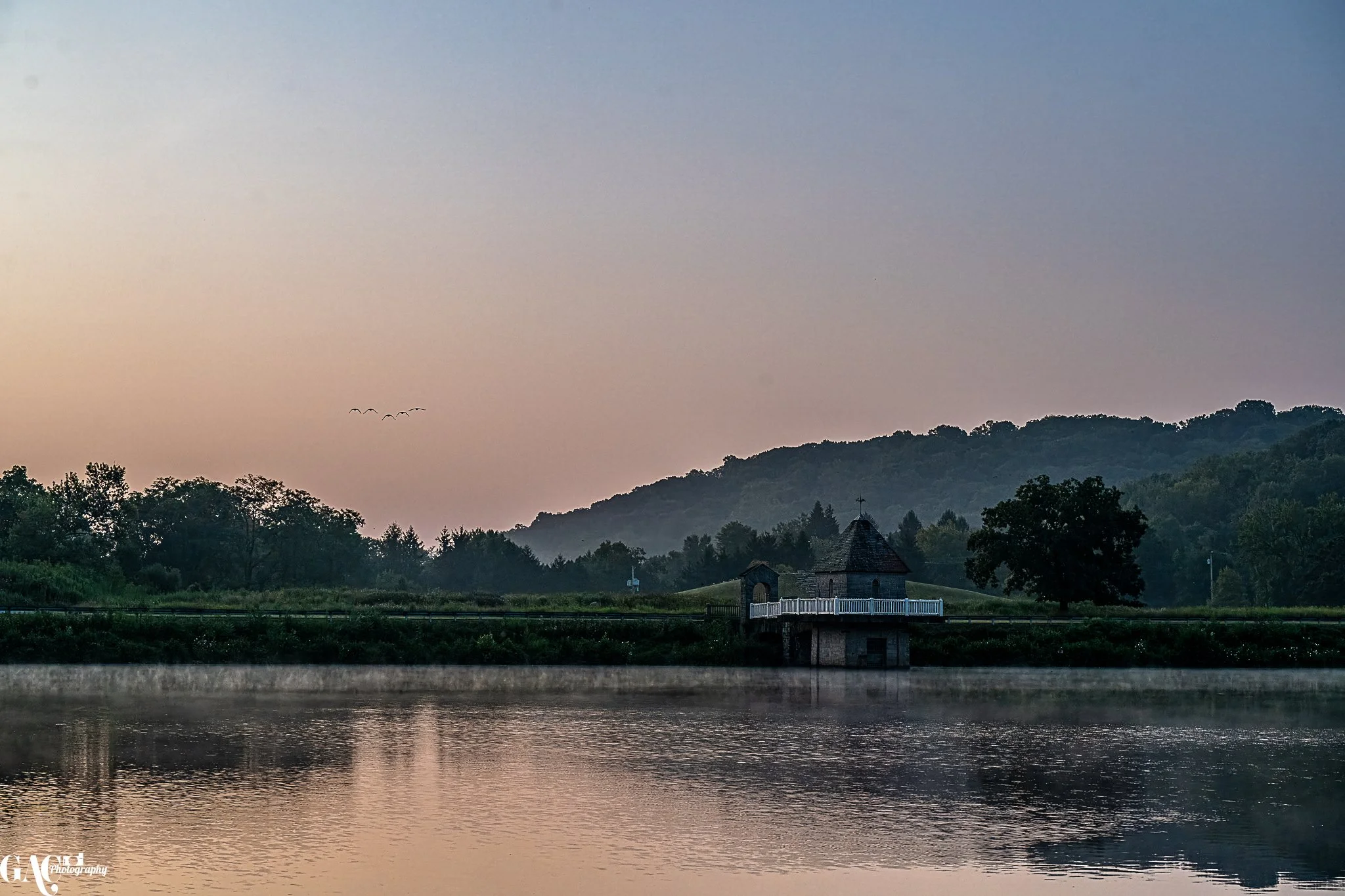 A calm lake with a stone building and white railing overlooking the water, surrounded by trees and hills at dusk.