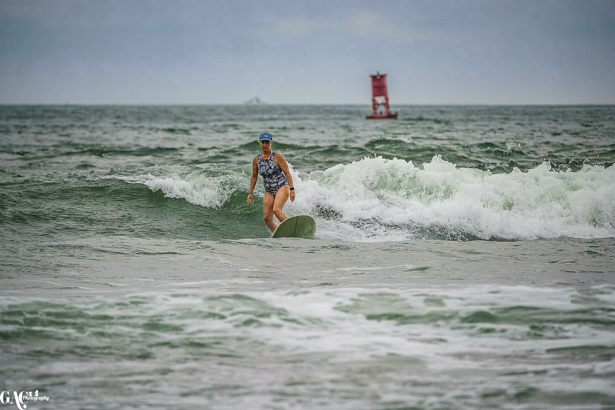 Woman surfing in the ocean with a buoy and overcast sky in the background.