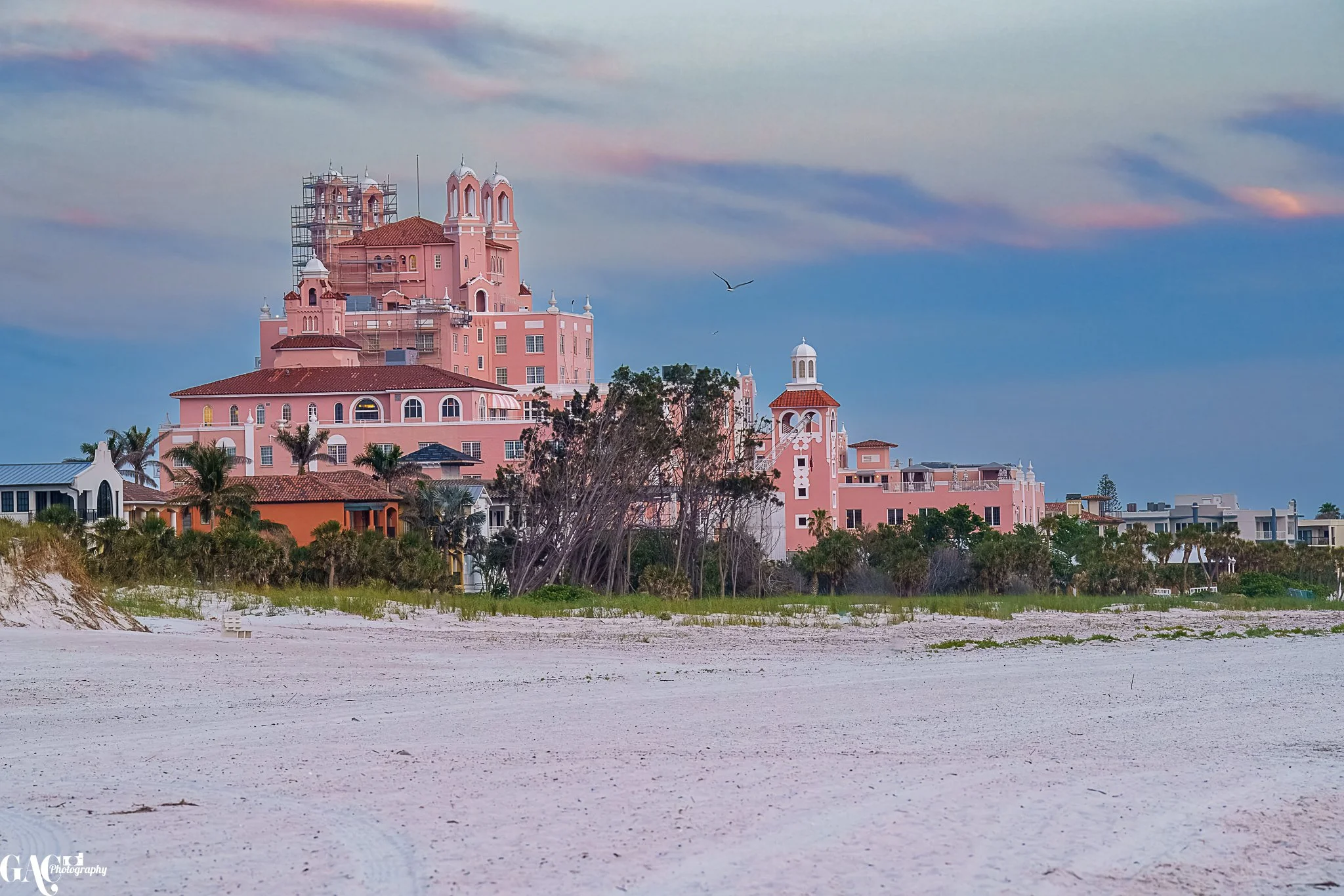 Large pink historic hotel with multiple towers and palm trees, located near a sandy beach under a gradient sky at sunset.