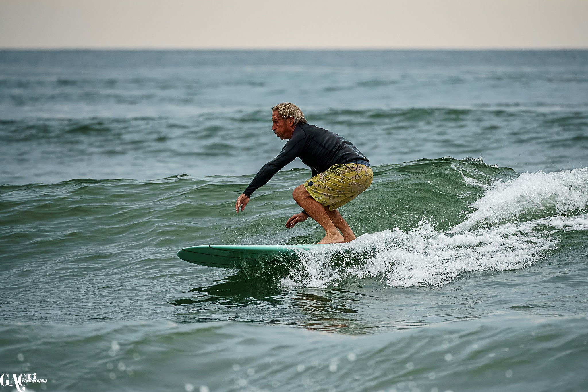 A man surfing on a green surfboard in the ocean, wearing a black long-sleeve rash guard and yellow swim shorts with a pattern.