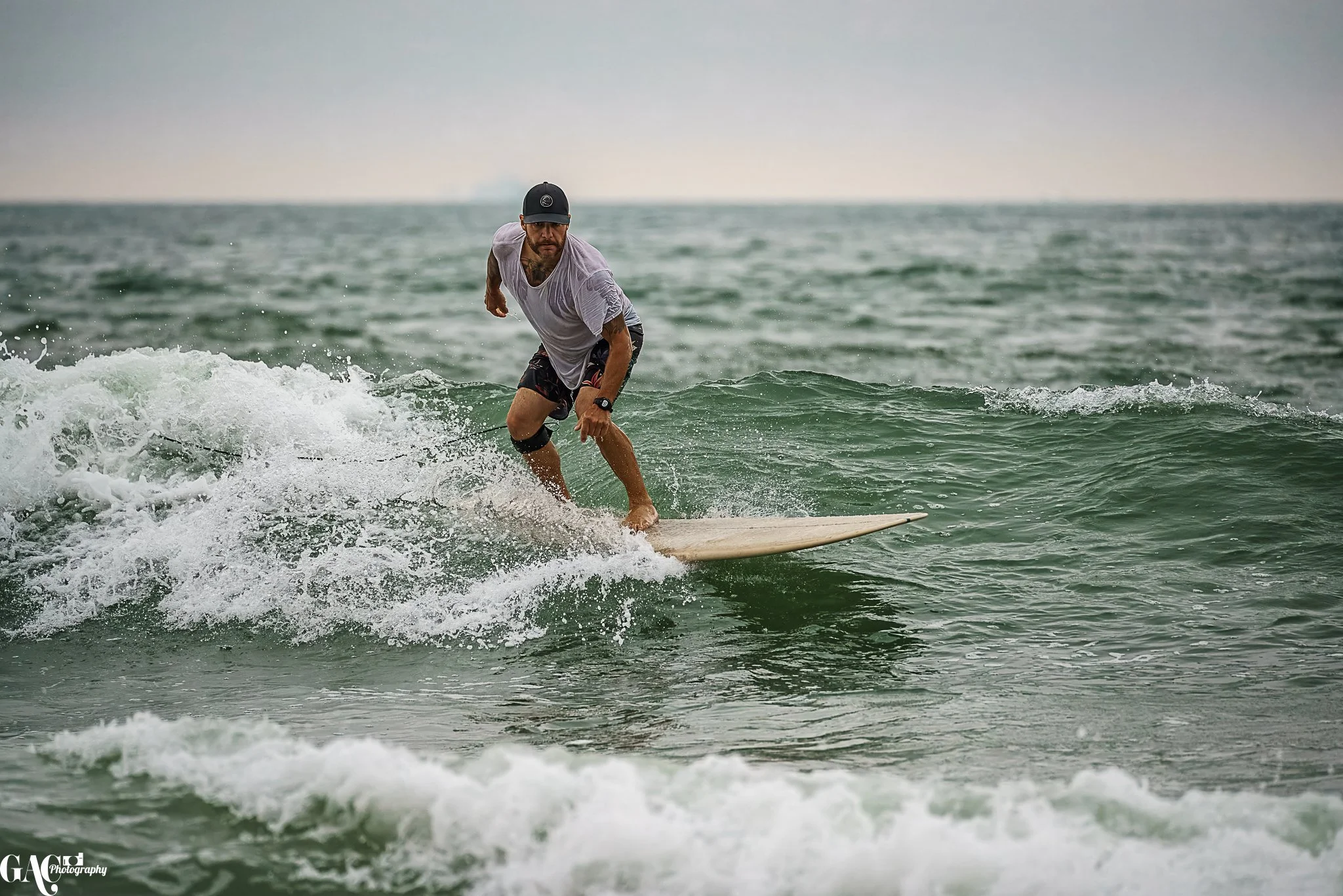 A man surfing on a small wave in the ocean, wearing a white t-shirt, black shorts, a black cap, and a smartwatch.