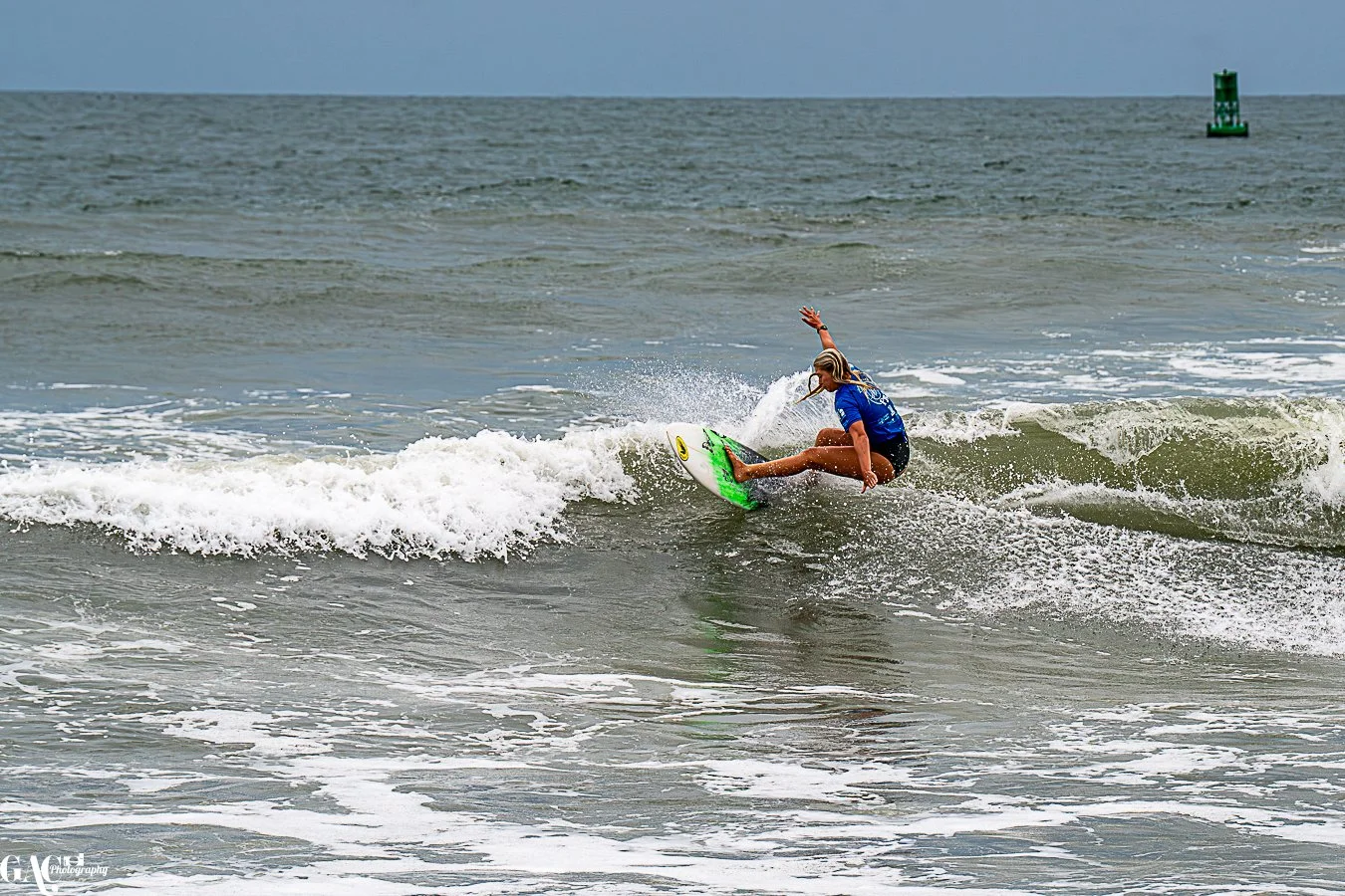 A woman surfing on a wave in the ocean, with a green buoy visible in the background.