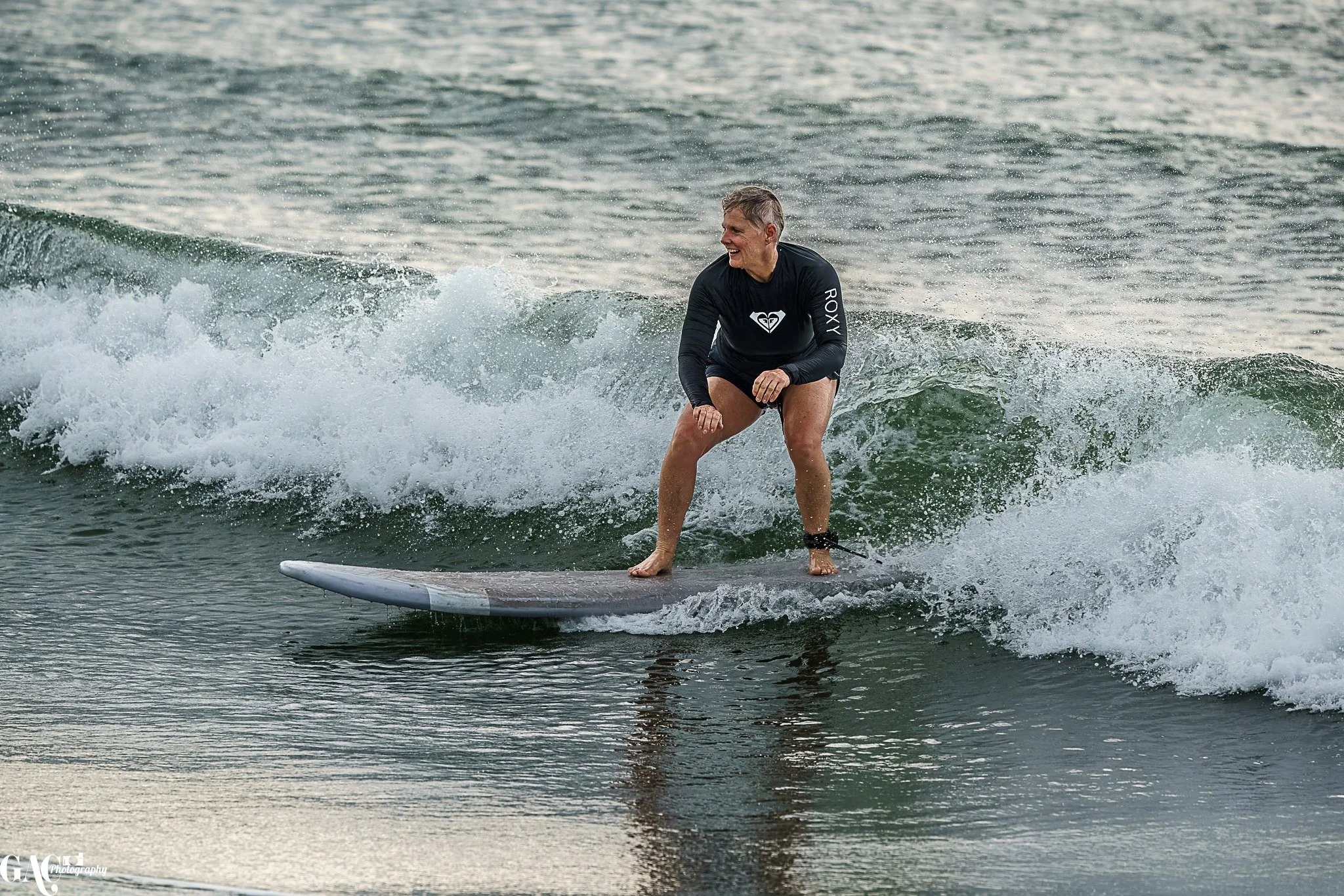 Woman surfing on a wave in the ocean, wearing a black wetsuit with brand logos, smiling and balancing on a surfboard.