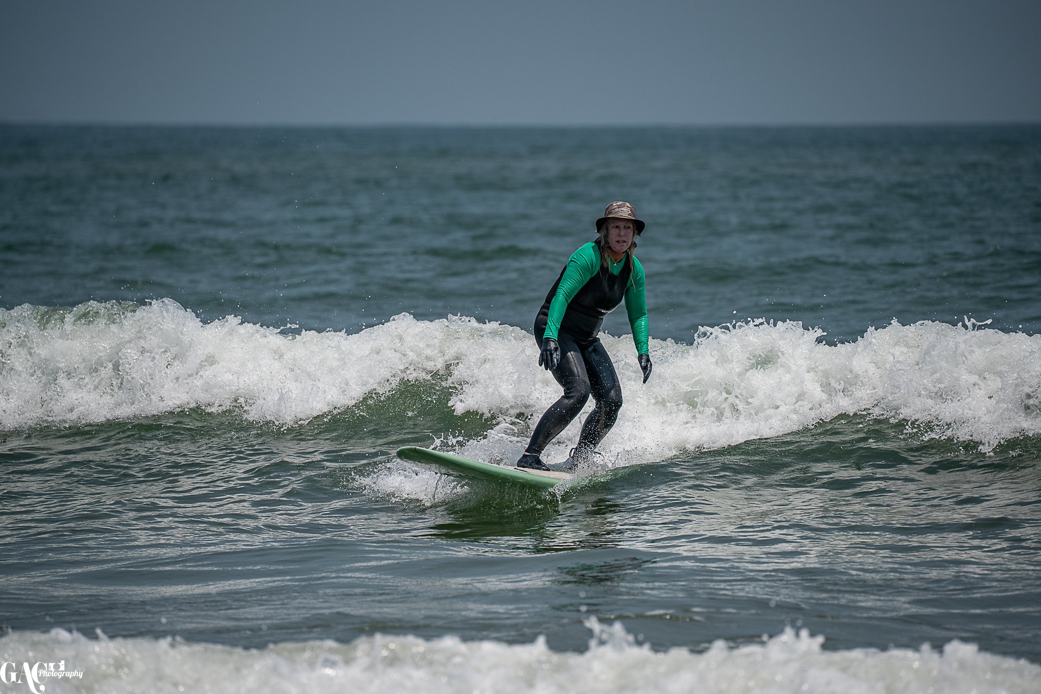 A woman wearing a hat, green long sleeve shirt, and black wetsuit riding a surfboard on small waves in the ocean.