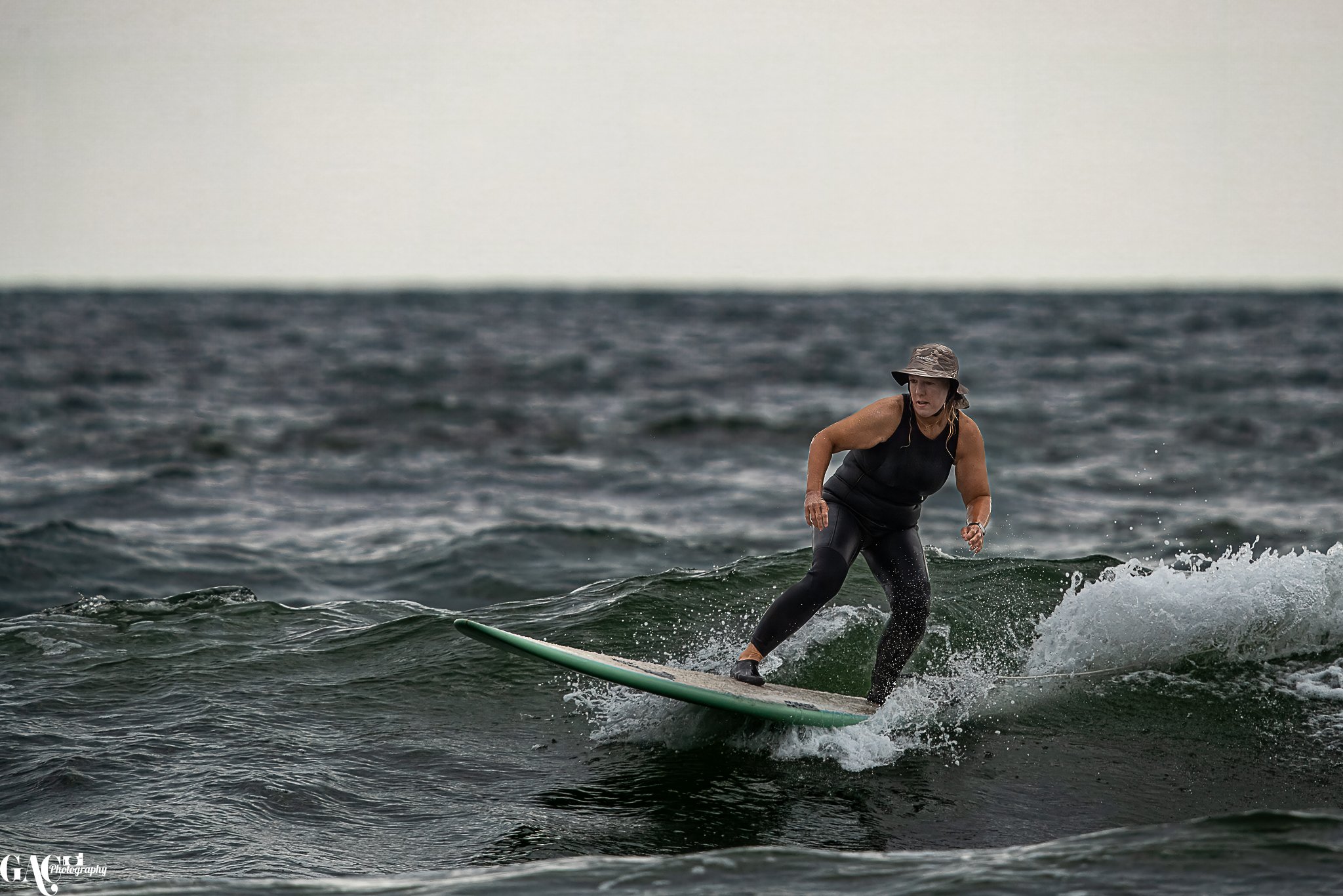 A woman surfing on a small wave in the ocean, wearing a hat and black wetsuit.