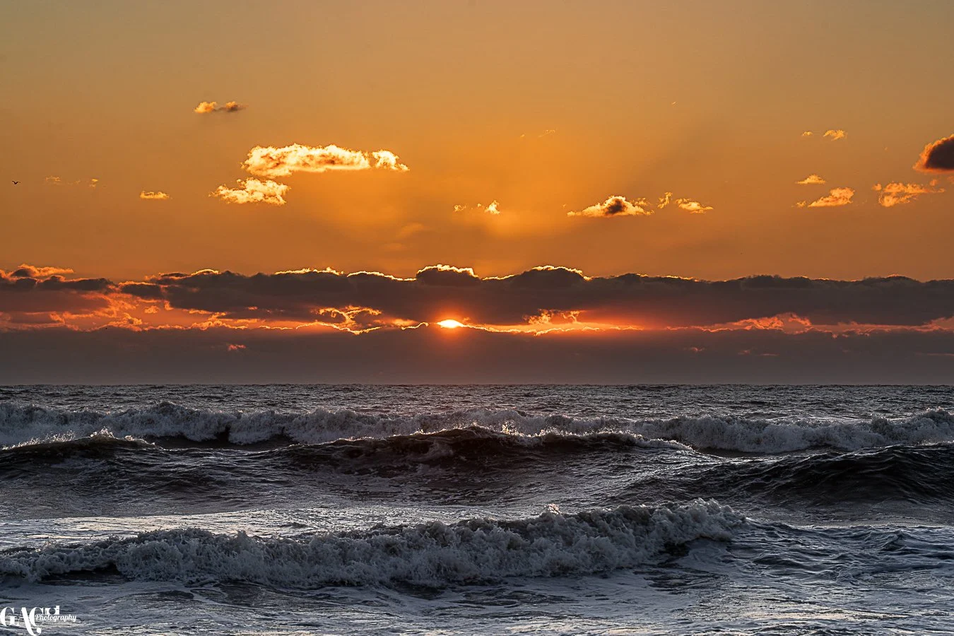 Sunset over the ocean with waves crashing and clouds in the sky.