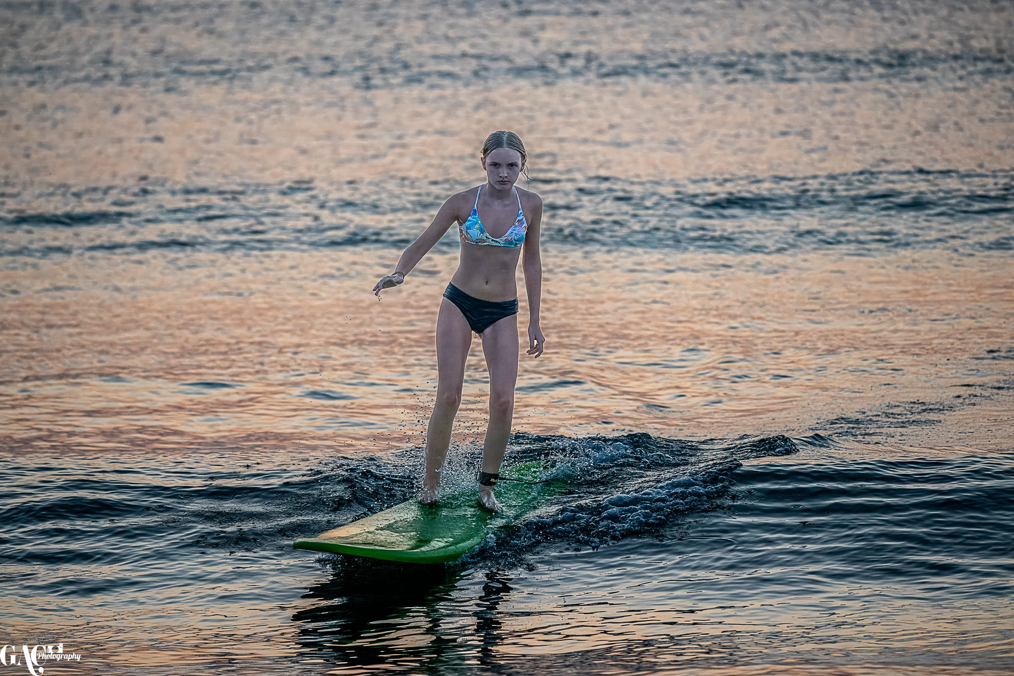 Young girl in a bikini riding a surfboard on calm water at sunset.