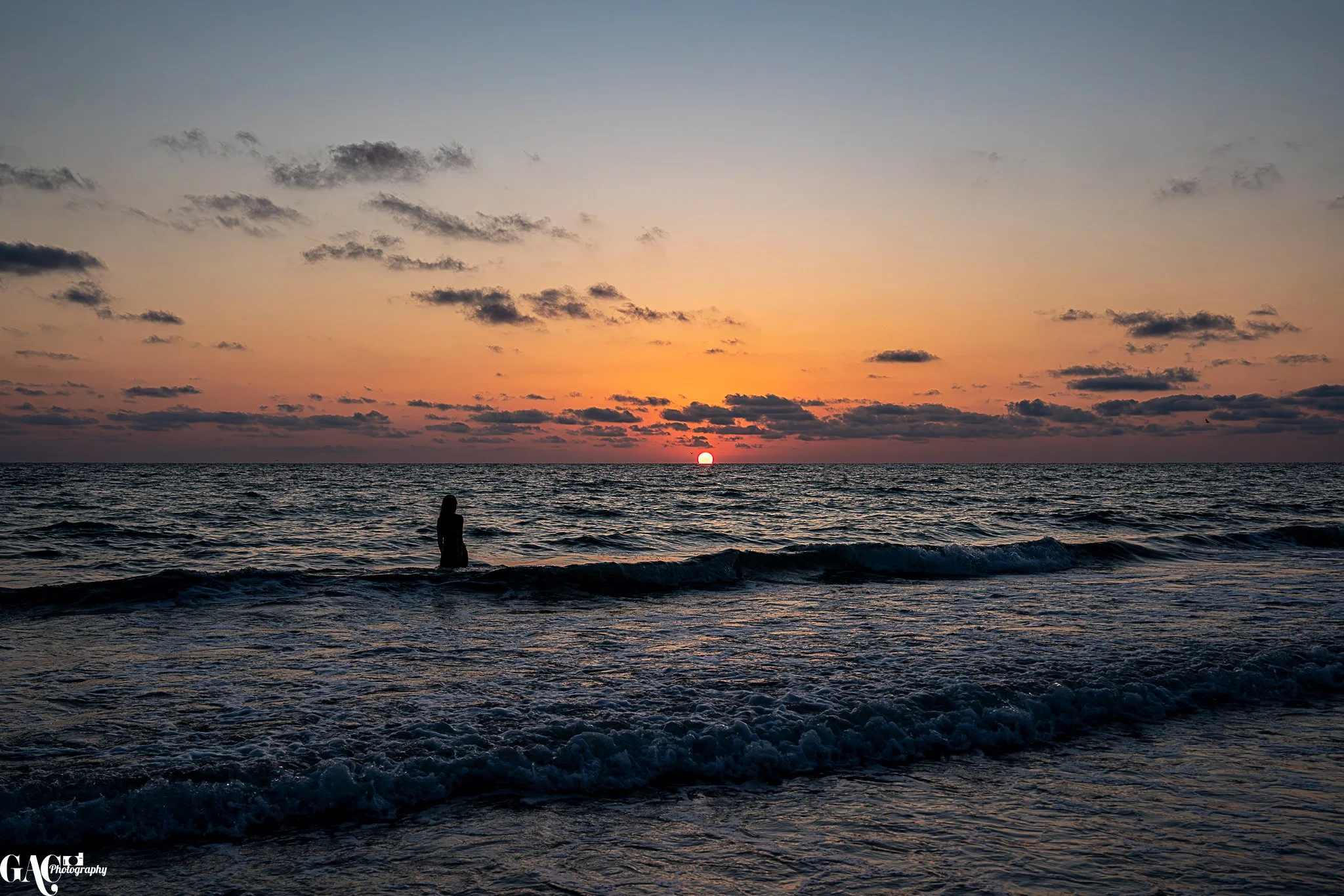 Beach sunset with a person standing in the ocean and clouds in the sky.