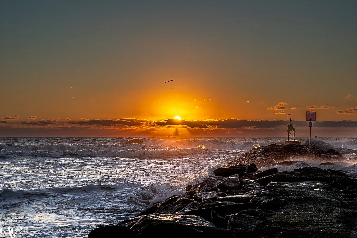 Sunset over the ocean with a rocky shoreline in the foreground, waves crashing, a bird flying in the sky, and a lighthouse on a small pier to the right.