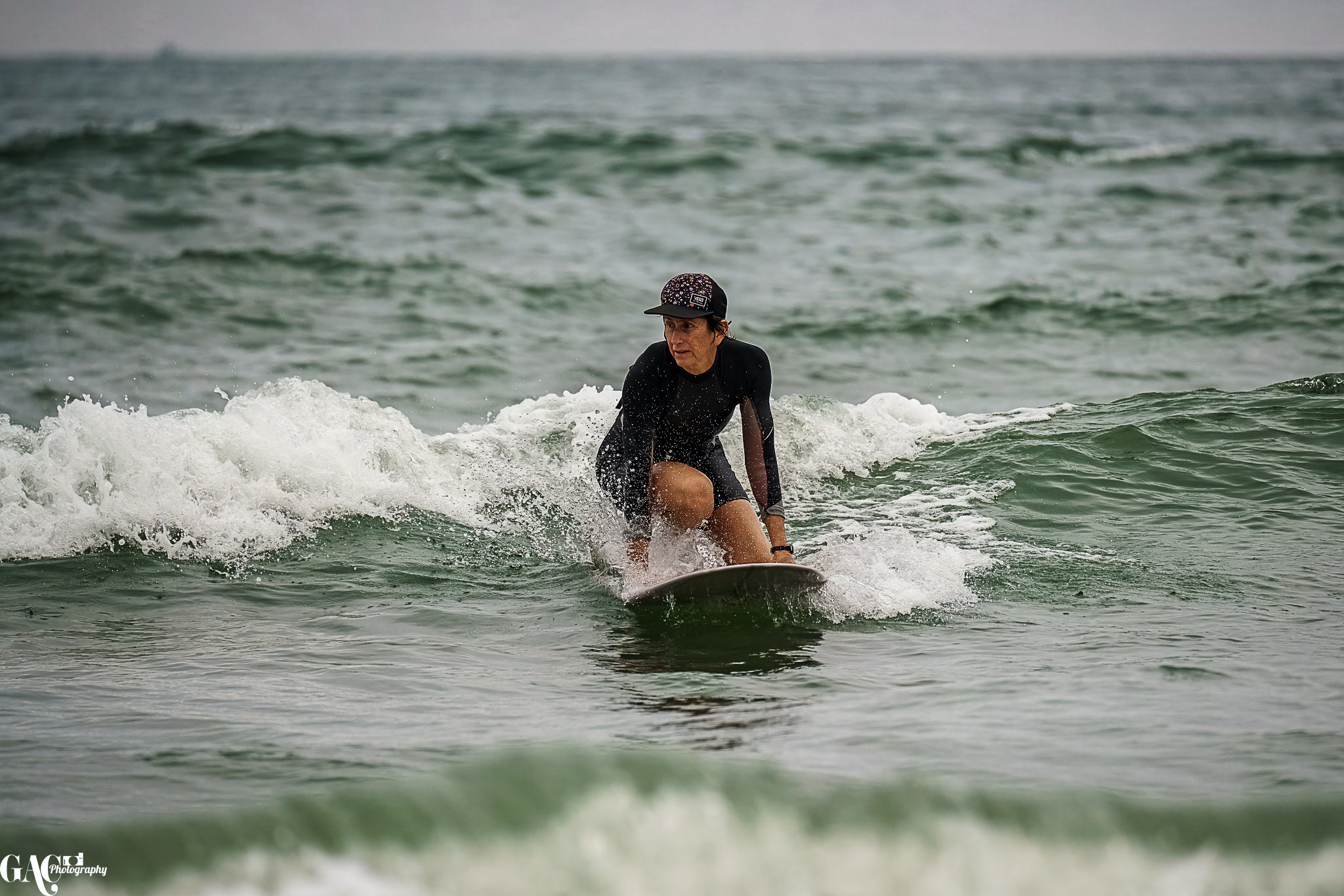 A woman surfing on a surfboard in the ocean, wearing a black wetsuit and a black cap.